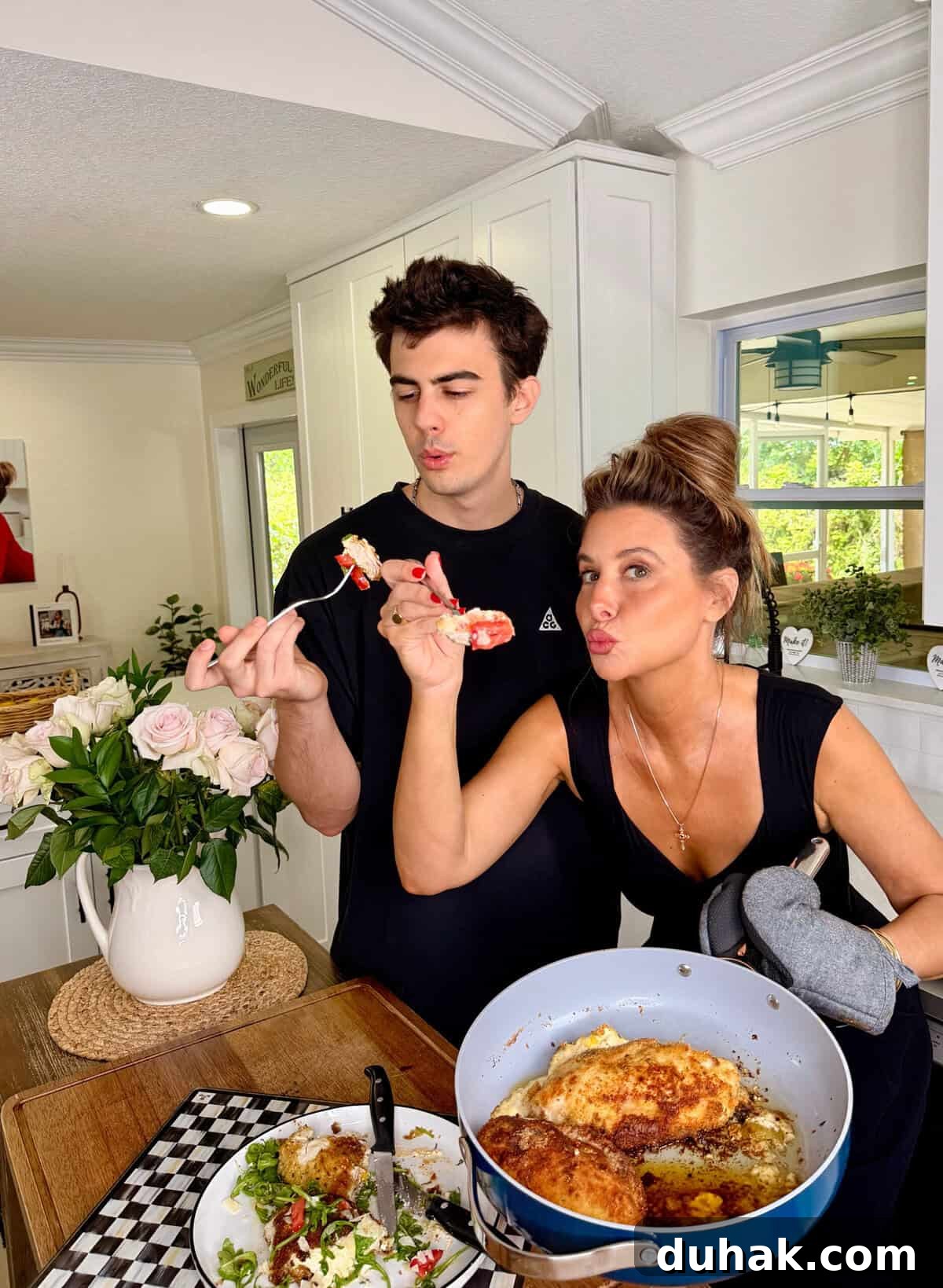 Creamy Spinach & Cheese Stuffed Chicken Cutlets Recipe 14 A young man and woman stand in a bright kitchen tasting food from a skillet. The woman, holding an oven mitt, feeds the man as they both make playful faces. A bouquet of roses is on the counter beside them.