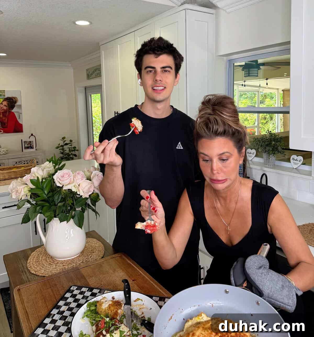 Creamy Spinach & Cheese Stuffed Chicken Cutlets Recipe 15 A young man and a woman stand in a bright kitchen, eating food. The woman holds a frying pan and wears an oven mitt. A vase of roses and a plate with food are on the table beside them. Both appear relaxed and casual.