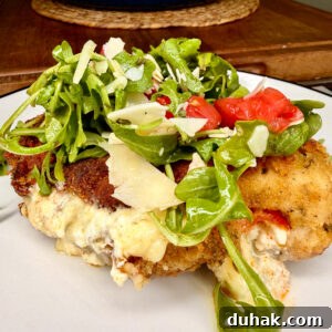 Creamy Spinach & Cheese Stuffed Chicken Cutlets Recipe 16 A breaded, cheese-stuffed chicken breast is topped with fresh arugula, sliced tomatoes, and shaved parmesan on a white plate, with a blue pot in the blurred background.