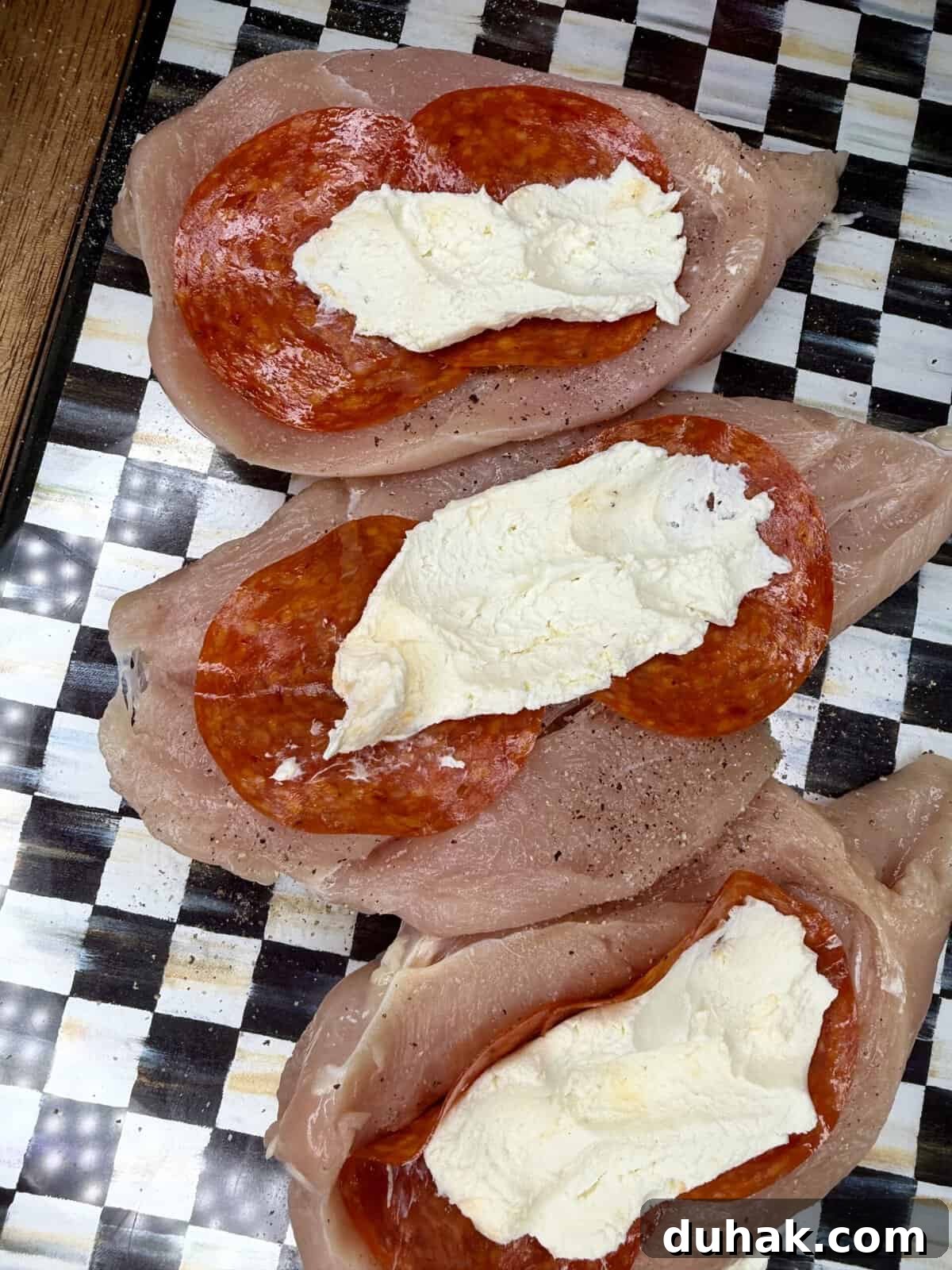 Creamy Spinach & Cheese Stuffed Chicken Cutlets Recipe 3 Three raw chicken breasts are topped with slices of pepperoni and a layer of creamy white cheese, arranged on a black-and-white checkered surface.