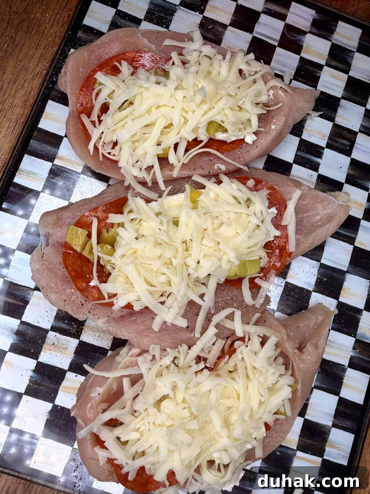 Creamy Spinach & Cheese Stuffed Chicken Cutlets Recipe 4 Three raw chicken breasts are sliced open and stuffed with slices of pepperoni, chopped green peppers, and shredded cheese, arranged on a black and white checkered surface.