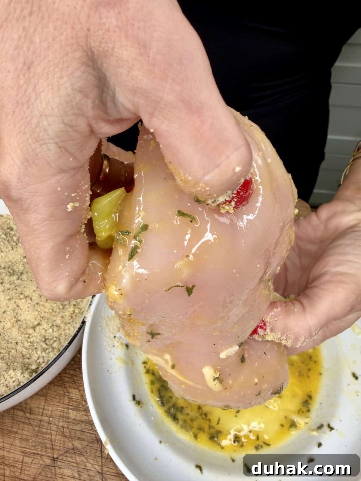 Creamy Spinach & Cheese Stuffed Chicken Cutlets Recipe 5 A person holds a raw chicken breast coated in egg wash and herbs above a bowl, preparing it for breading; a plate of breadcrumbs is visible nearby.