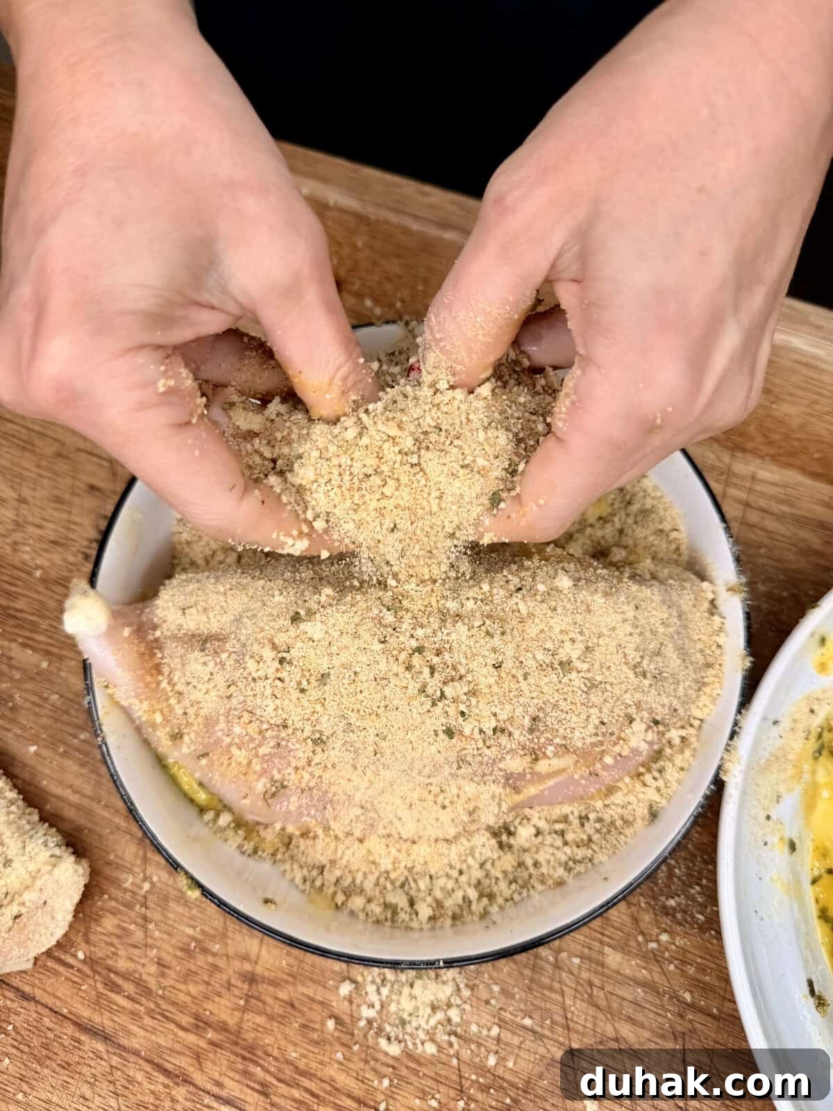 Creamy Spinach & Cheese Stuffed Chicken Cutlets Recipe 6 Two hands coat a raw chicken leg with seasoned breadcrumbs in a shallow bowl on a wooden surface, preparing it for cooking.