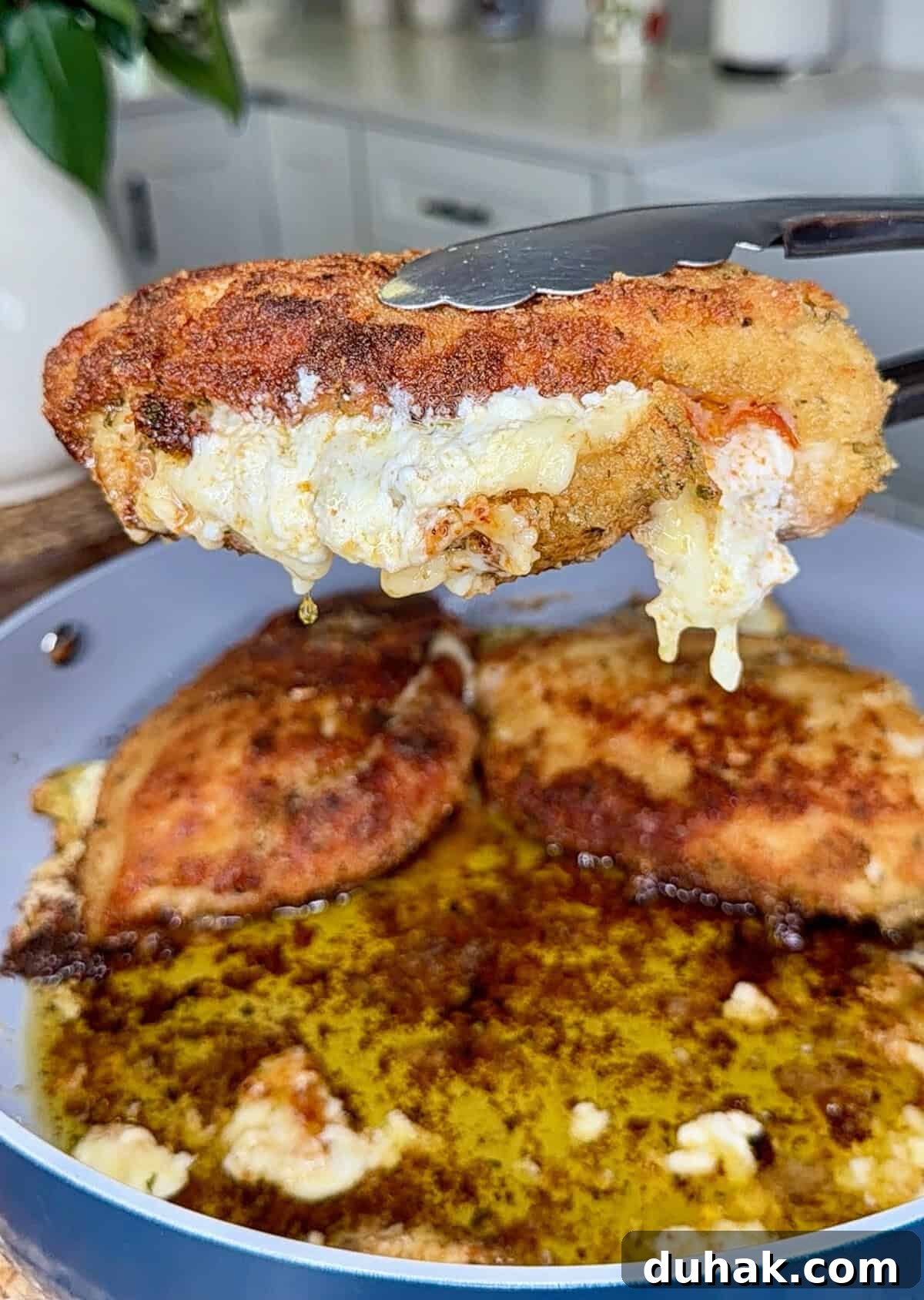 Creamy Spinach & Cheese Stuffed Chicken Cutlets Recipe 9 A close-up of tongs holding a breaded, fried chicken breast with melted cheese oozing out, above a pan with more pieces cooking in bubbling oil. The kitchen background is slightly blurred.