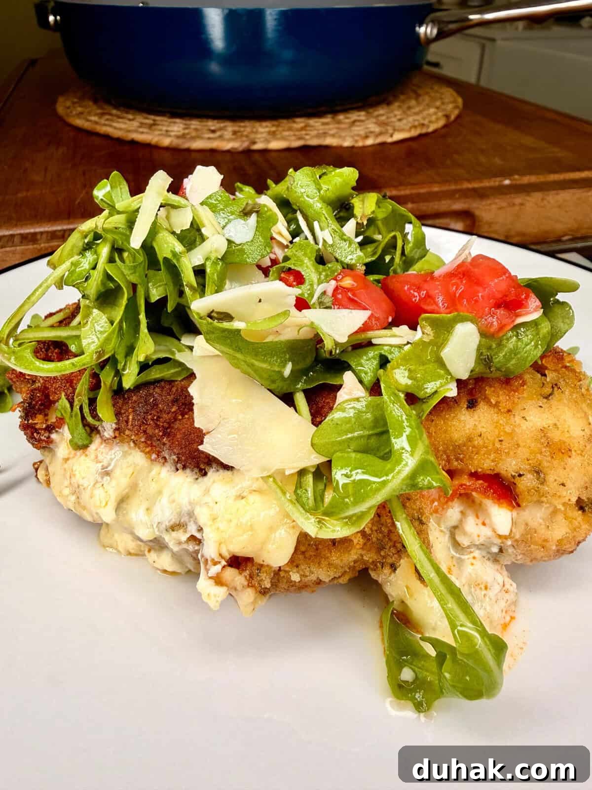 Creamy Spinach & Cheese Stuffed Chicken Cutlets Recipe 10 A breaded, stuffed chicken breast topped with a fresh arugula salad, shaved parmesan, and chopped tomatoes sits on a white plate, with a pot and cutting board in the background.