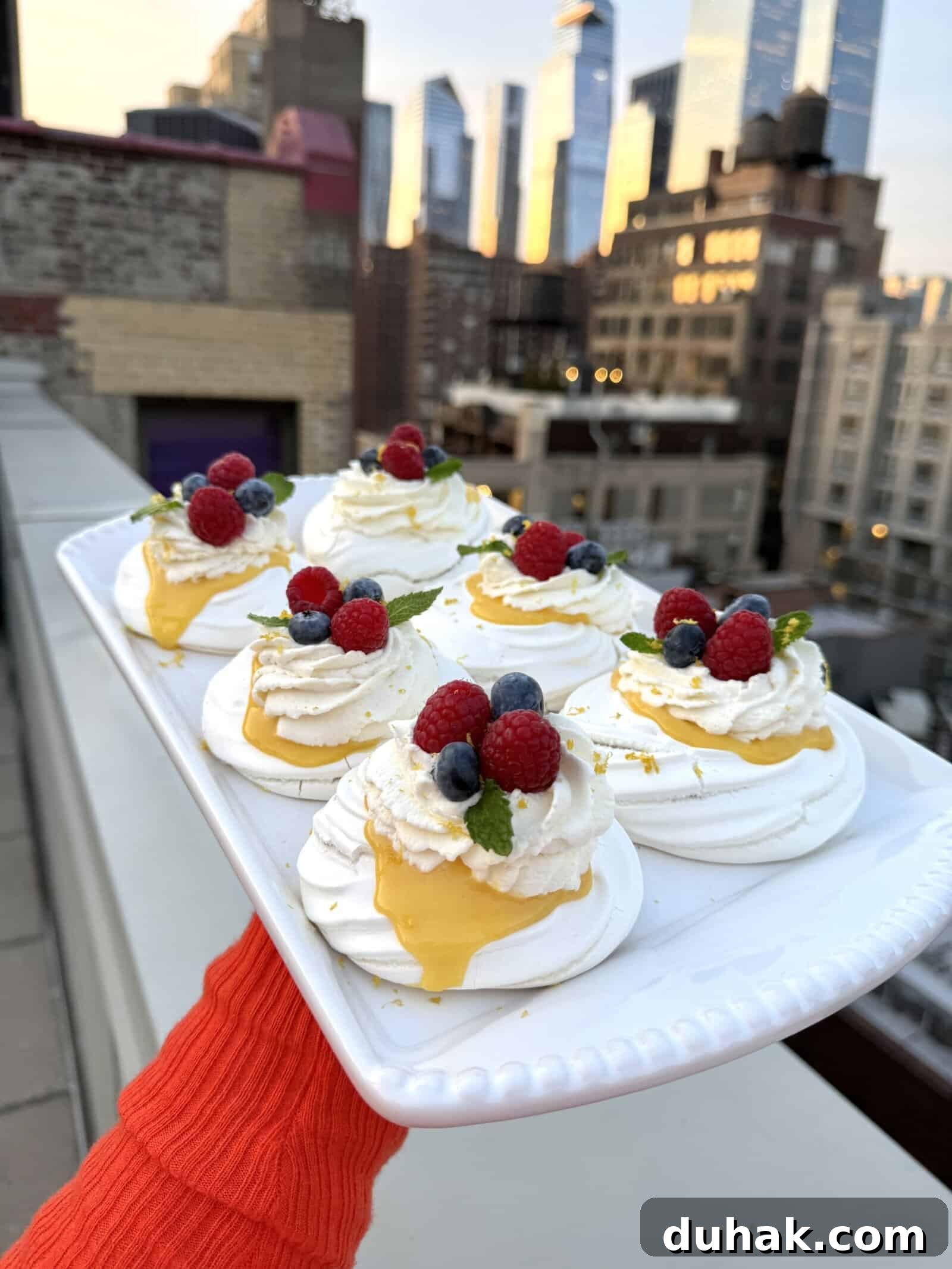 Yuzu Curd Pavlova Recipe: Light Citrus Meringue Cake 2 A person holds a rectangular platter of mini pavlovas topped with whipped cream, lemon curd, raspberries, blueberries, and mint on a rooftop, with a city skyline in the background at sunset.
