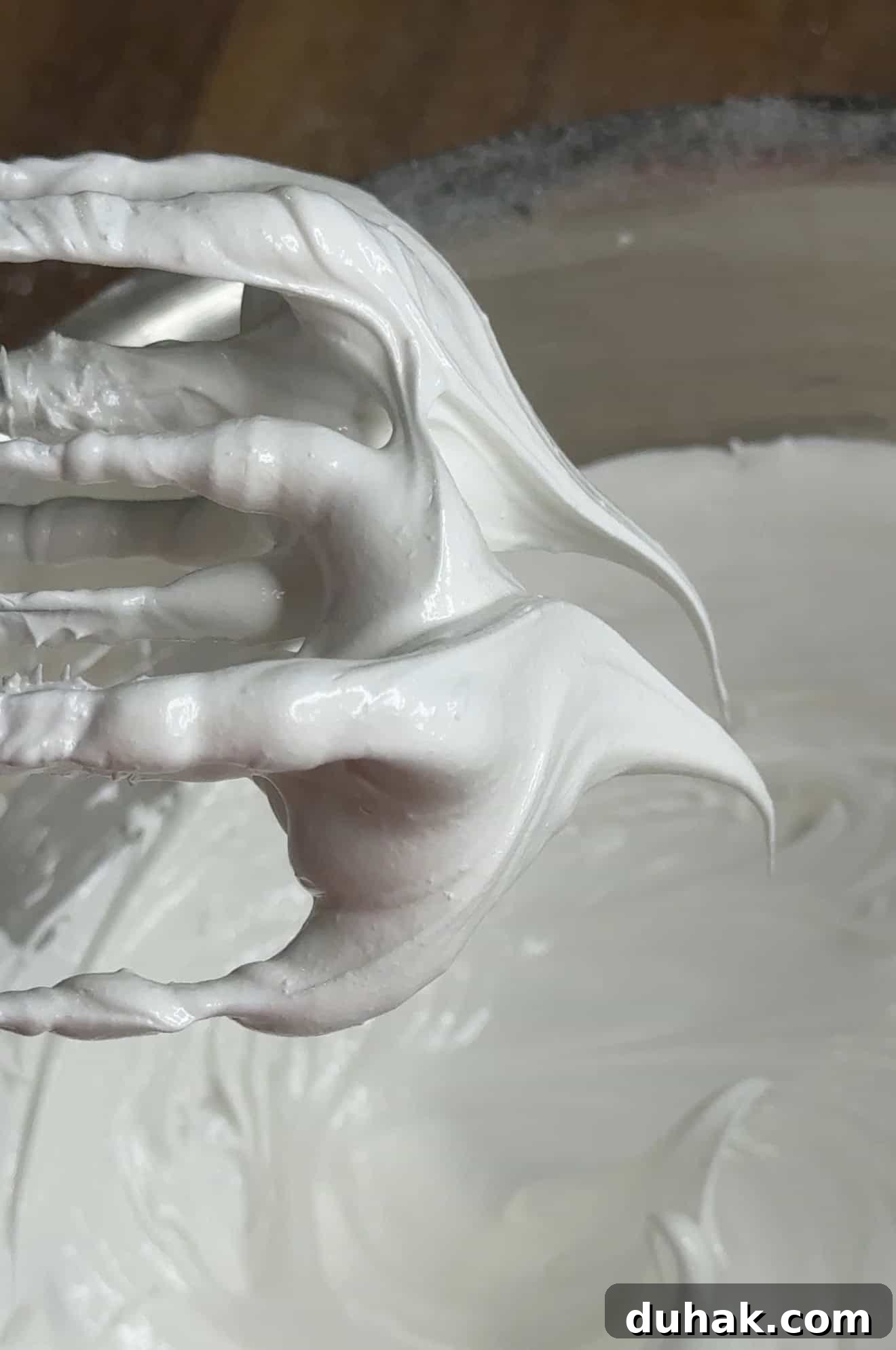 Yuzu Curd Pavlova Recipe: Light Citrus Meringue Cake 3 A close-up of a whisk lifted from a bowl, covered in glossy, white meringue with stiff peaks forming on the whisk and in the bowl below.