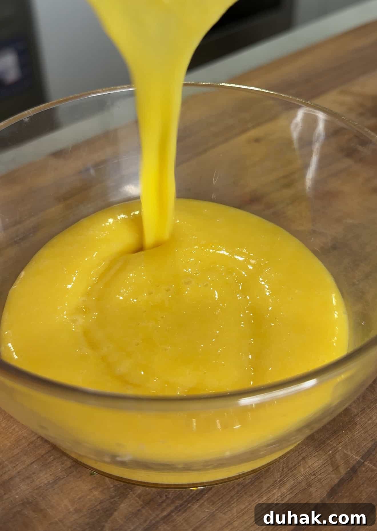 Yuzu Curd Pavlova Recipe: Light Citrus Meringue Cake 5 A thick, yellow liquid is being poured into a clear glass bowl, creating a smooth swirl on a wooden surface.