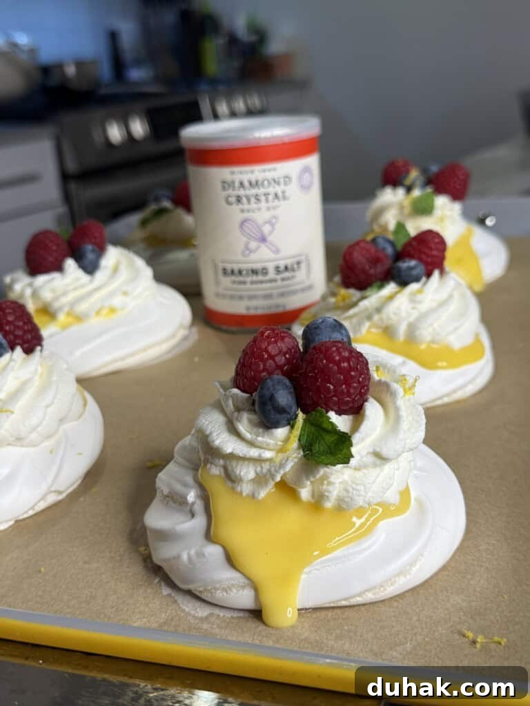 Yuzu Curd Pavlova Recipe: Light Citrus Meringue Cake 6 Mini pavlovas topped with whipped cream, raspberries, blueberries, mint leaves, and lemon curd sit on parchment paper. A container of Diamond Crystal baking salt is in the background on a kitchen counter.