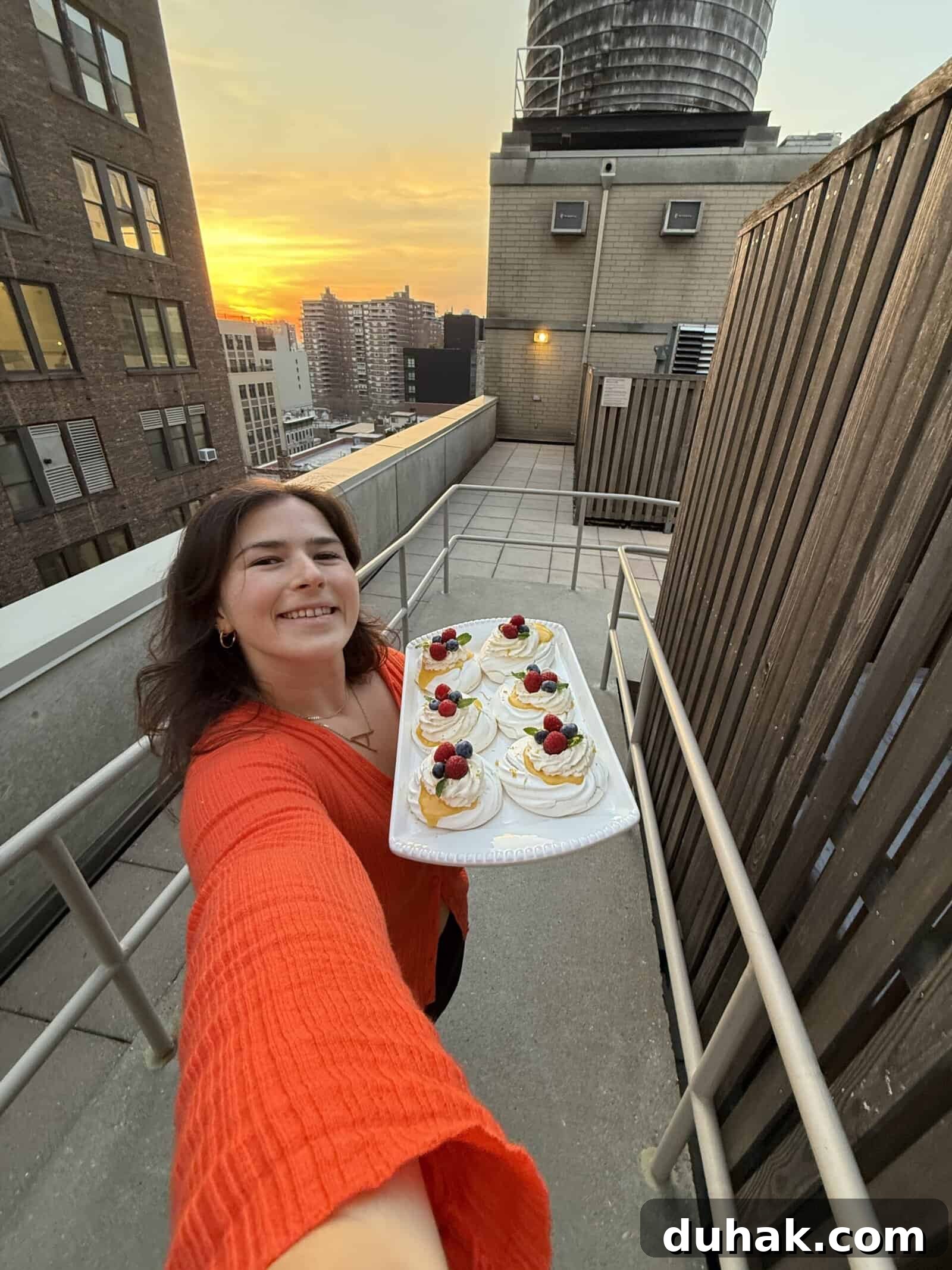 Yuzu Curd Pavlova Recipe: Light Citrus Meringue Cake 7 A woman in an orange top smiles and holds a tray of pavlova topped with berries on a rooftop at sunset, with tall city buildings in the background.