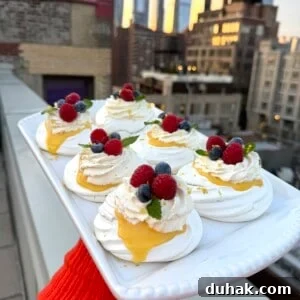 Yuzu Curd Pavlova Recipe: Light Citrus Meringue Cake 8 A person holds a rectangular platter of mini pavlovas topped with whipped cream, lemon curd, raspberries, blueberries, and mint on a rooftop, with a city skyline in the background at sunset.