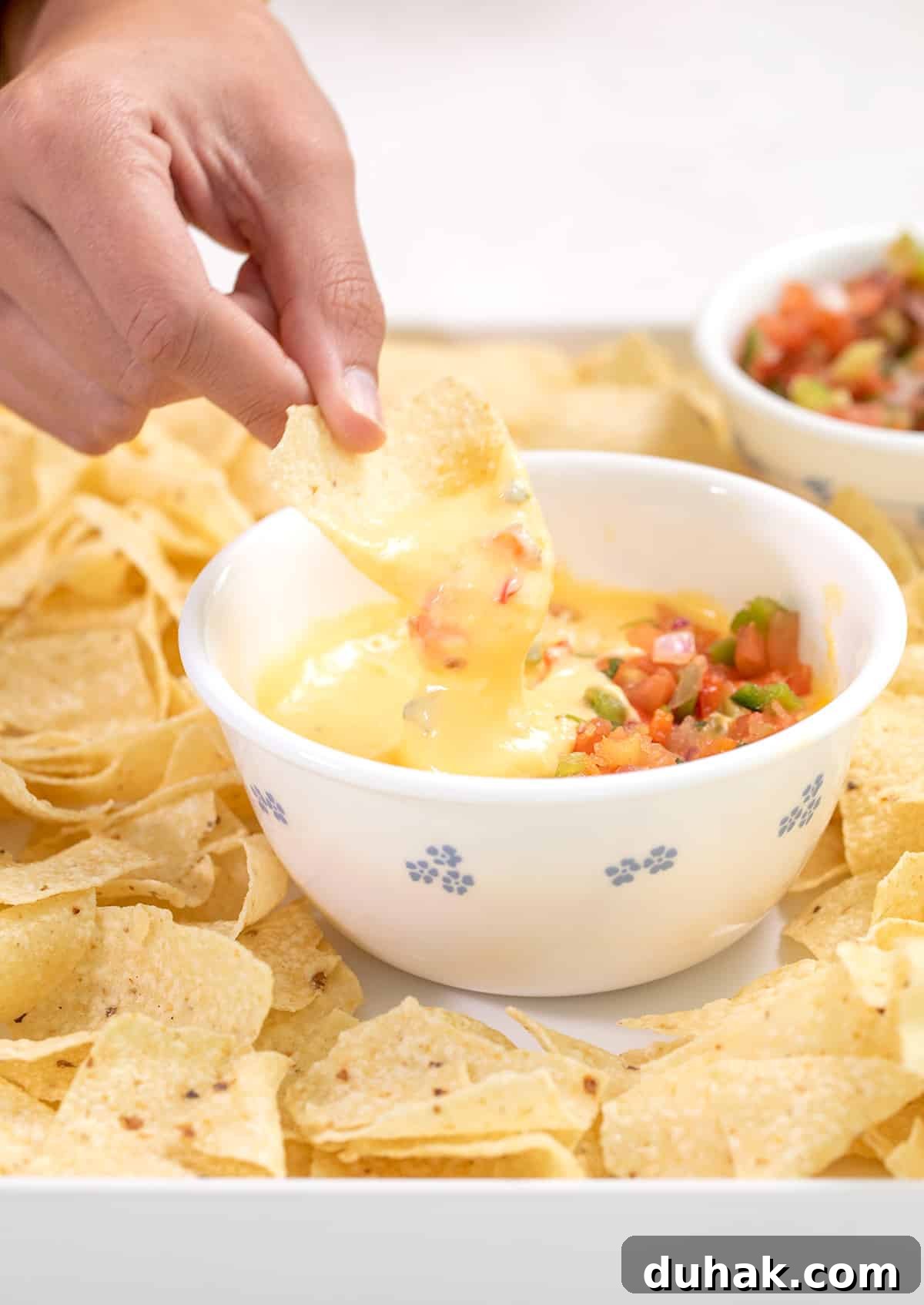 Hand dipping a chip in cheese sauce in a small white bowl with salsa on top