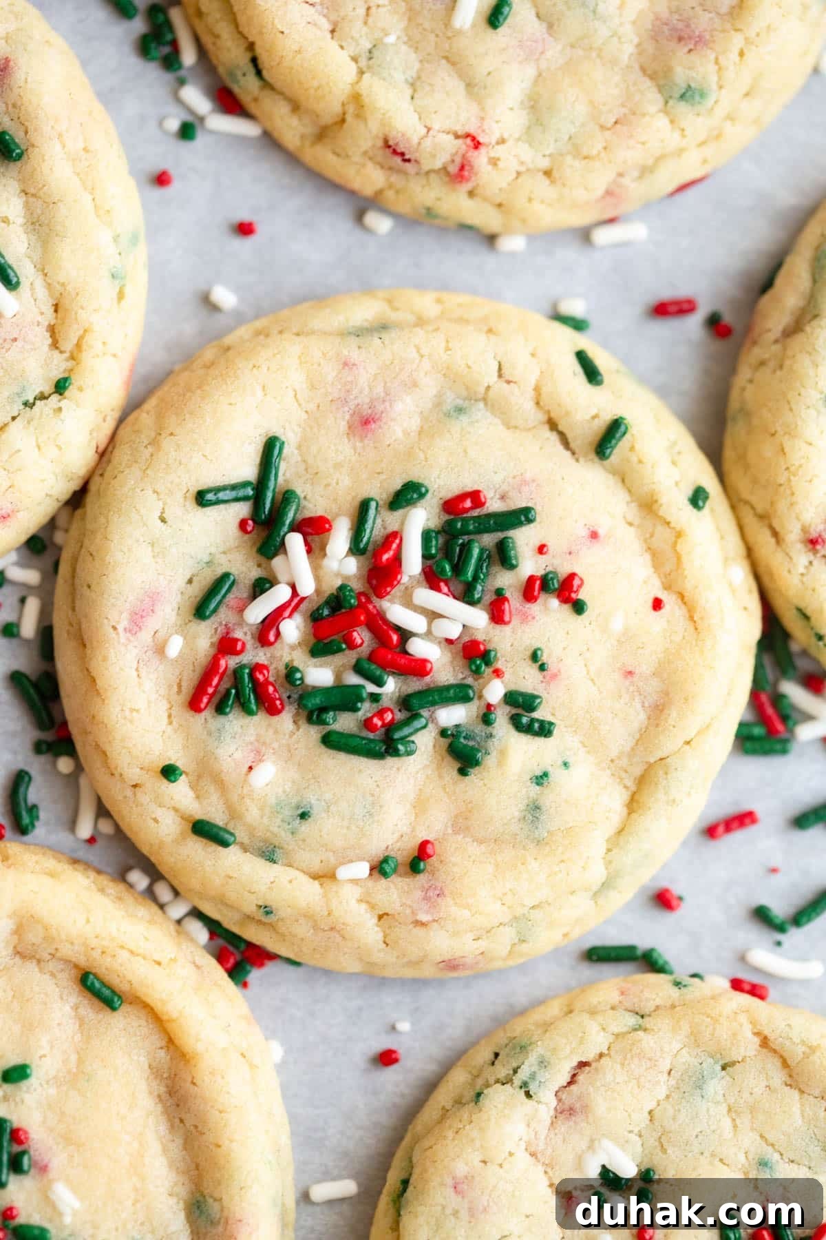 Christmas Sprinkle Sugar Cookies Recipe for Festive Holiday Baking 2 Christmas Sprinkle Sugar Cookies on a baking sheet lined with parchment paper and additional sprinkles scattered around them.