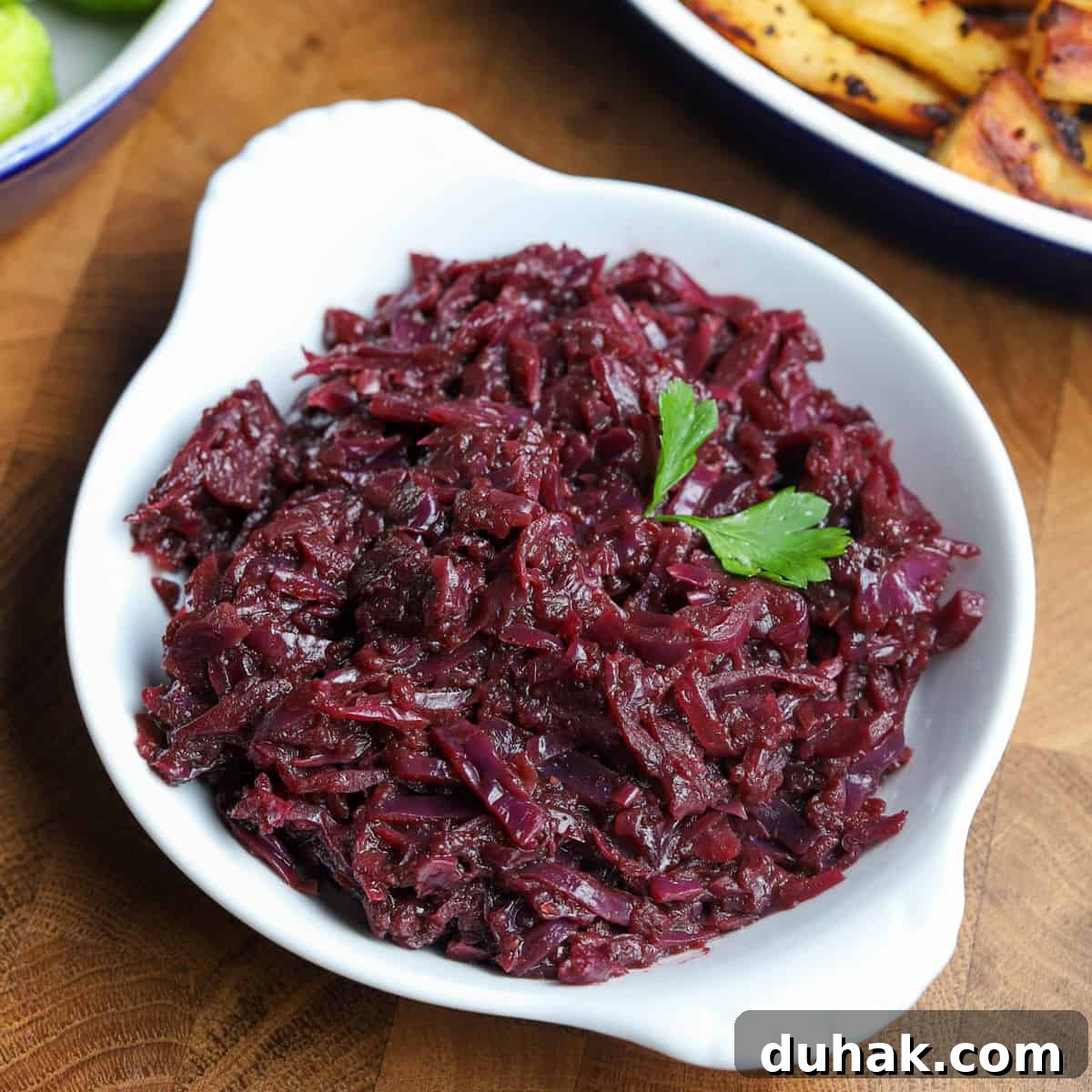 Photo of a white dish filled with braised red cabbage on a wooden table top with parsnips and sprouts in the background.