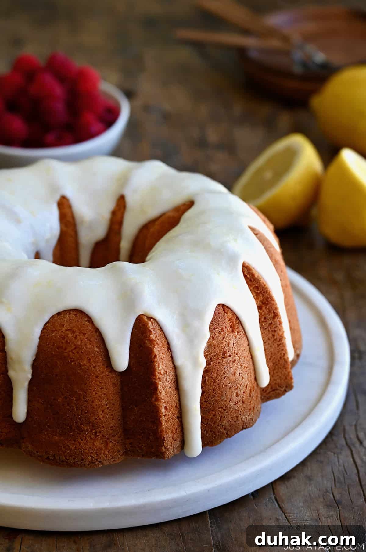 Glazed Lemon Pound Cake Recipe with Zesty Drizzle 2 A glazed lemon pound cake on a round, white serving platter with a bowl filled with raspberries in the background.