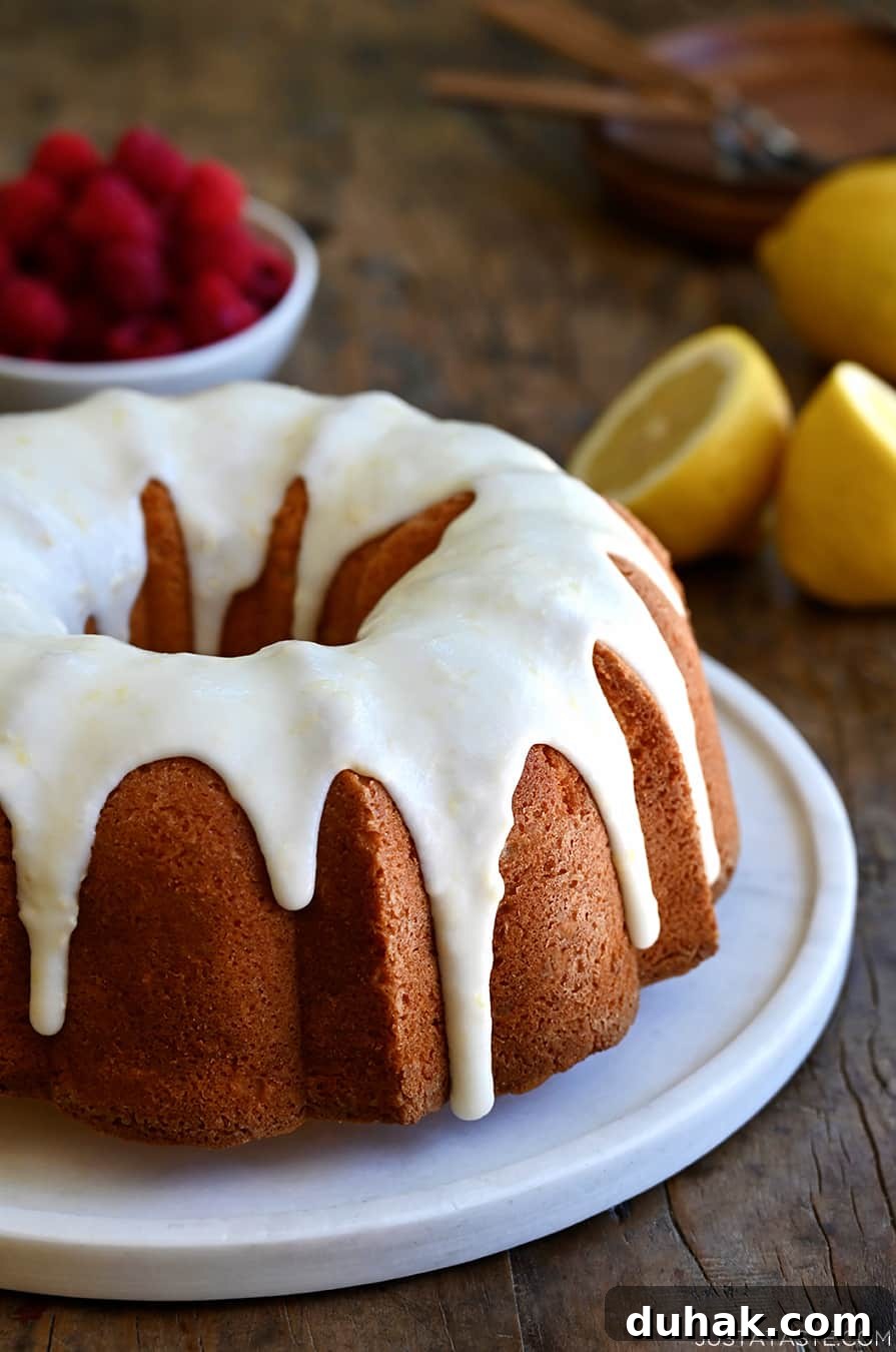 Glazed Lemon Pound Cake Recipe with Zesty Drizzle 10 A glazed lemon pound cake on a round, white serving platter with a bowl filled with raspberries in the background