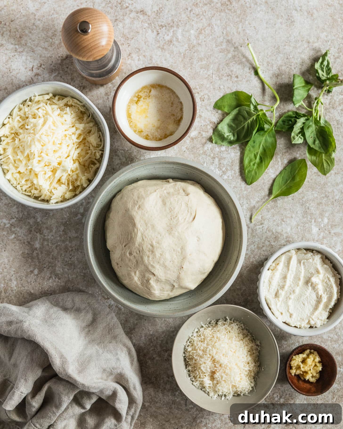 Cacio e Pepe Pizza Recipe: Crisp Crust with Creamy Pepper Sauce 3 White and wood bowls of pizza dough, mozzarella, Parmesan, ricotta, butter, and garlic next to basil on a tan counter.