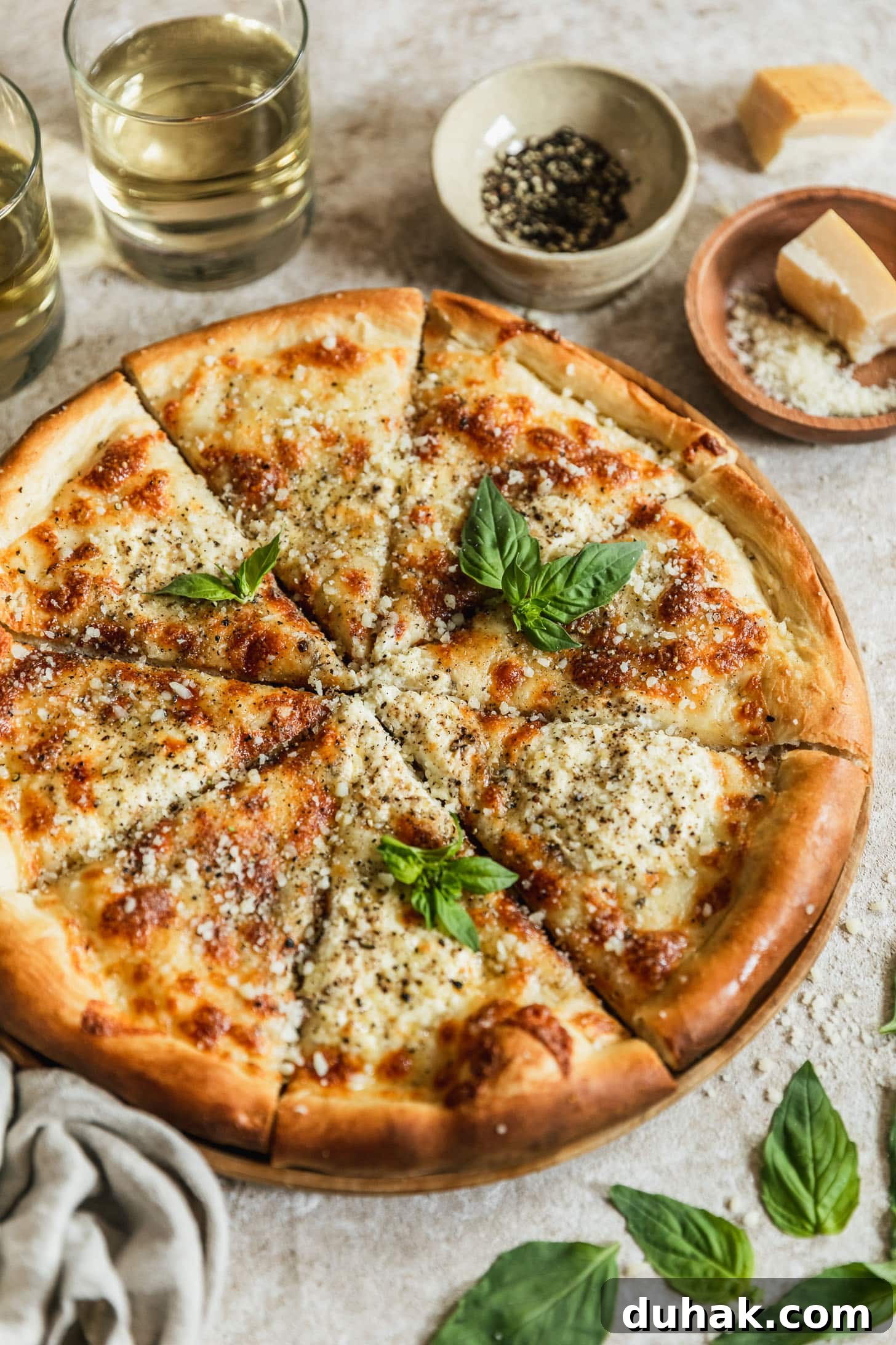 Cacio e Pepe Pizza Recipe: Crisp Crust with Creamy Pepper Sauce 6 A wood board with a cacio e pepe pizza on a beige counter next to fresh basil, two glasses of white wine, a white bowl of pepper, and a wood bowl of Pecorino Romano.