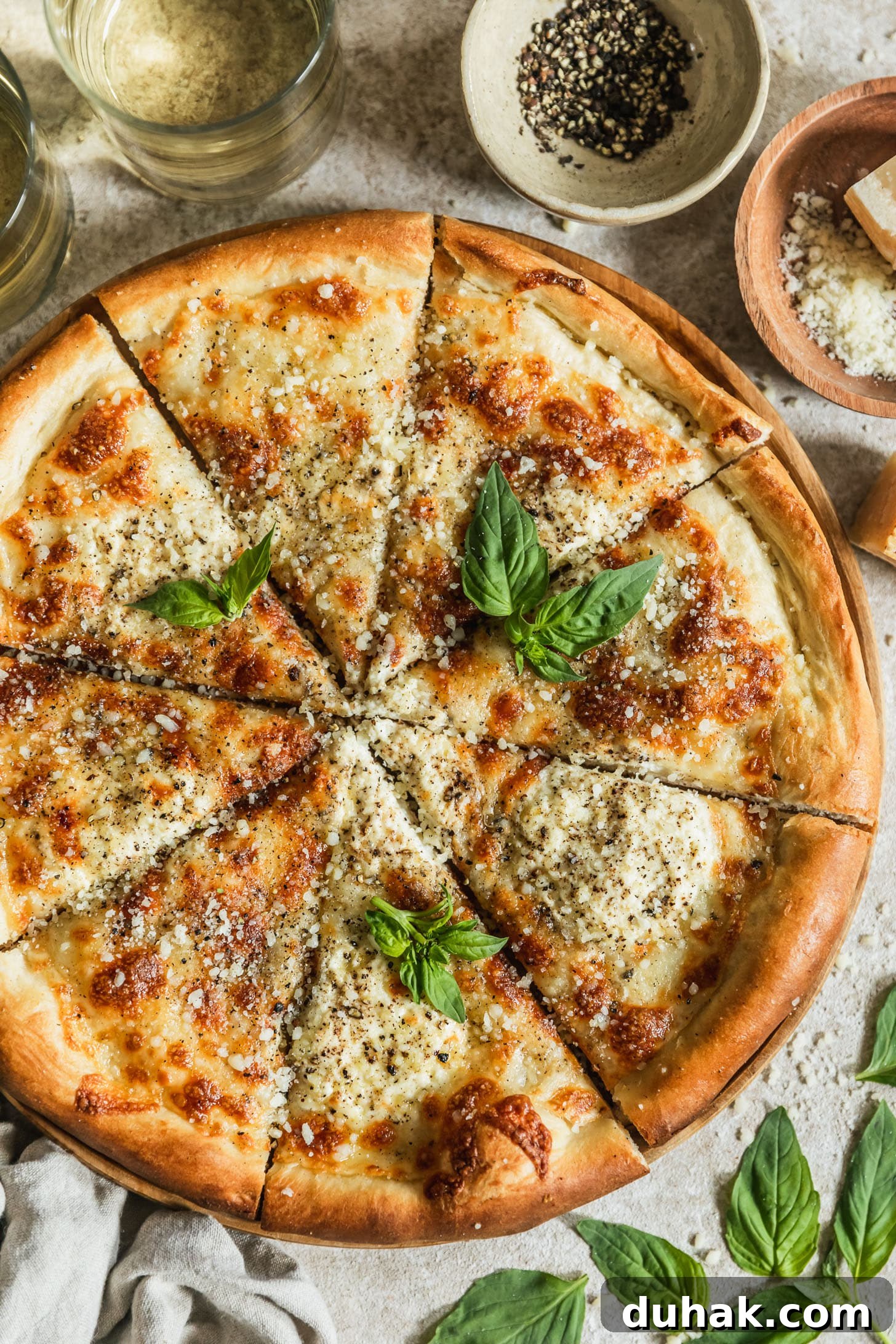 Cacio e Pepe Pizza Recipe: Crisp Crust with Creamy Pepper Sauce 7 Sliced cacio e pepe pizza with basil on a wood board next to basil leaves, a beige linen, two glasses of white wine, a white bowl of pepper, and a wood bowl of Pecorino Romano on a beige counter.