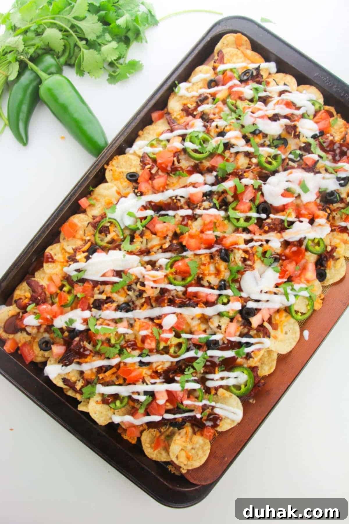 A tray of loaded nachos with jalapeños, tomatoes, black olives, and a drizzle of sour cream on a white background.