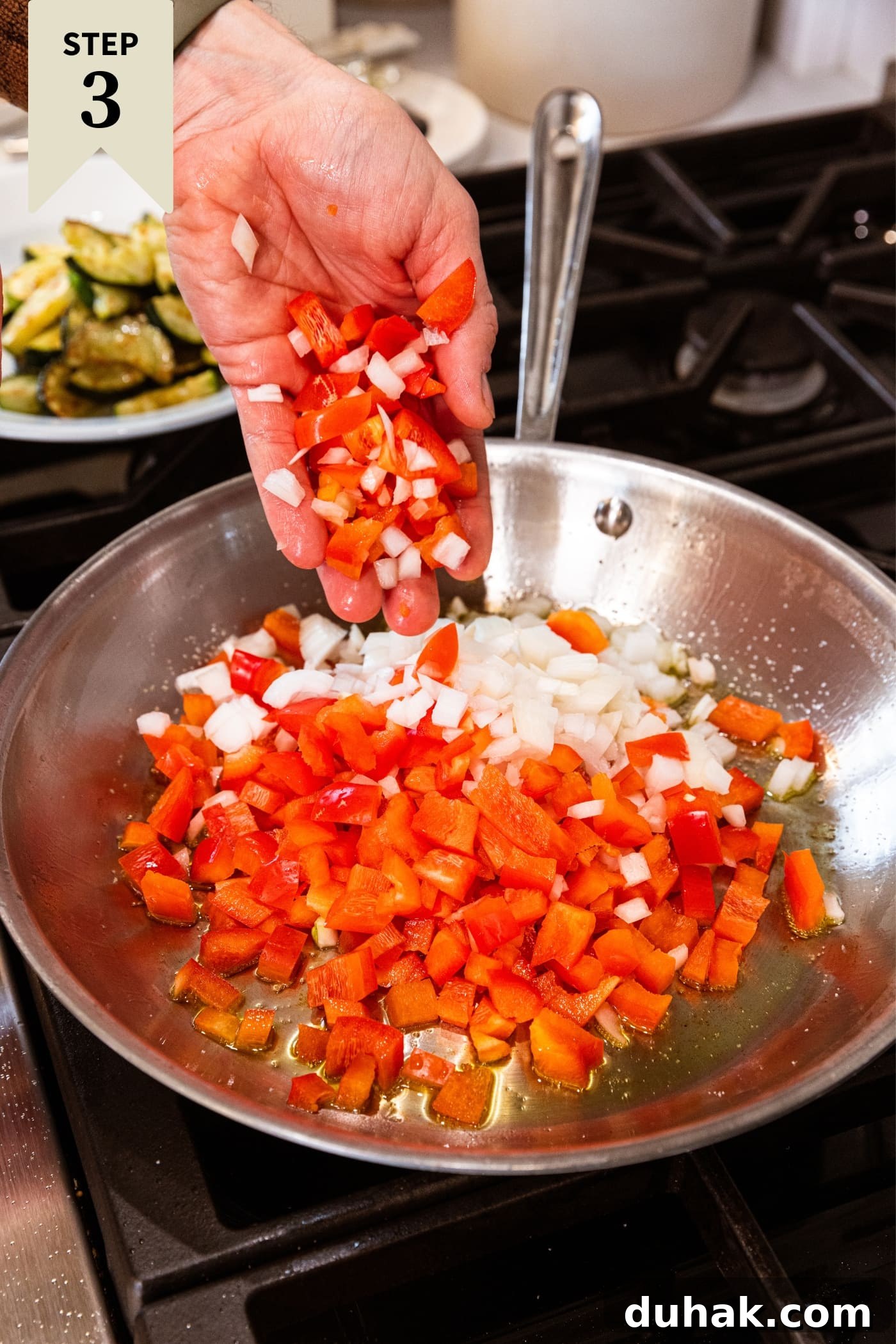 Cheesy Turkey & Spaghetti Squash Casserole Recipe 11 Hand dropping diced red bell pepper and onion into a metal skillet on the stovetop with hot oil inside with cooked zucchini in background.