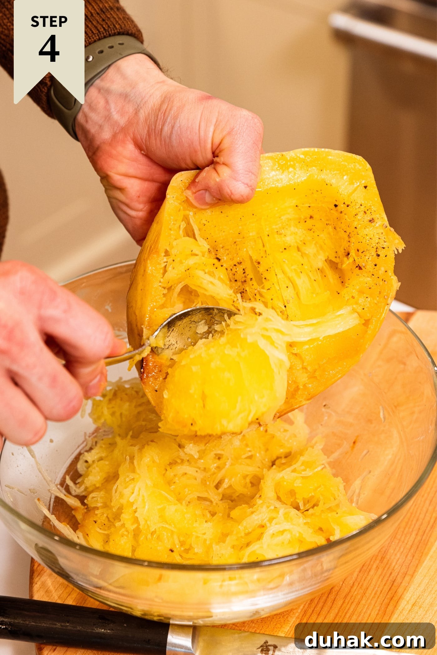 Cheesy Turkey & Spaghetti Squash Casserole Recipe 15 Hand holding roasted spaghetti squash scooping out the soft interior into a large glass bowl on wood cutting board.