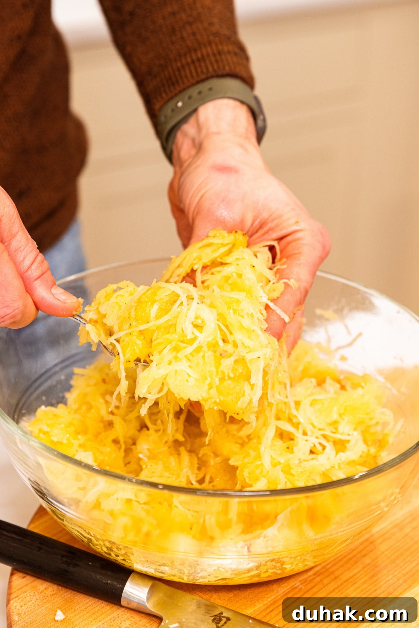 Cheesy Turkey & Spaghetti Squash Casserole Recipe 16 Hand using spoon and other hand to break up the texture of yellow-colored spaghetti squash in large glass bowl.