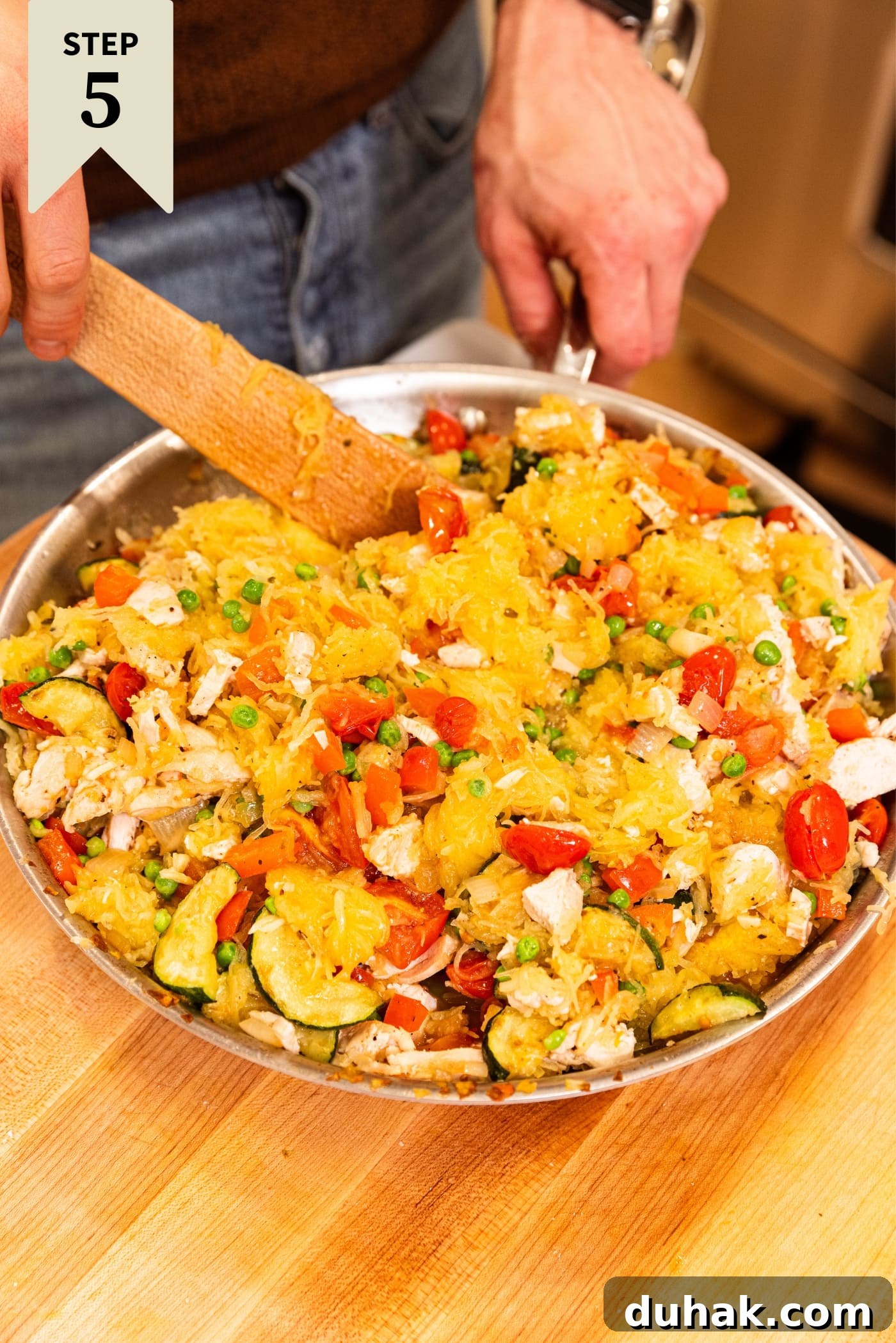 Cheesy Turkey & Spaghetti Squash Casserole Recipe 17 Metal skillet with tomatoes, spaghetti squash, chicken, and more vegetables inside being mixed together with wooden spatula on wooden surface.