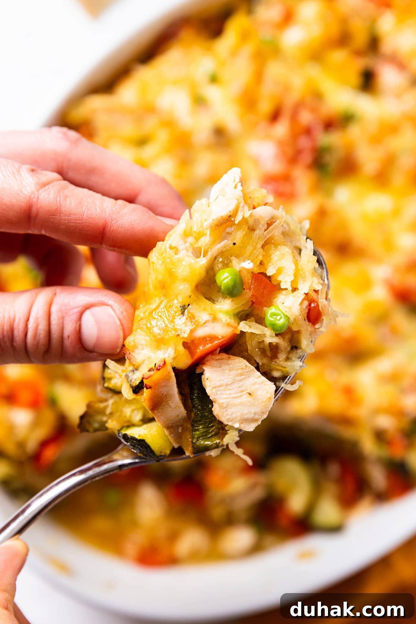 Cheesy Turkey & Spaghetti Squash Casserole Recipe 21 Hand holding spoonful of spaghetti squash chicken casserole with all ingredients showing including zucchini, peas, squash, and chicken.
