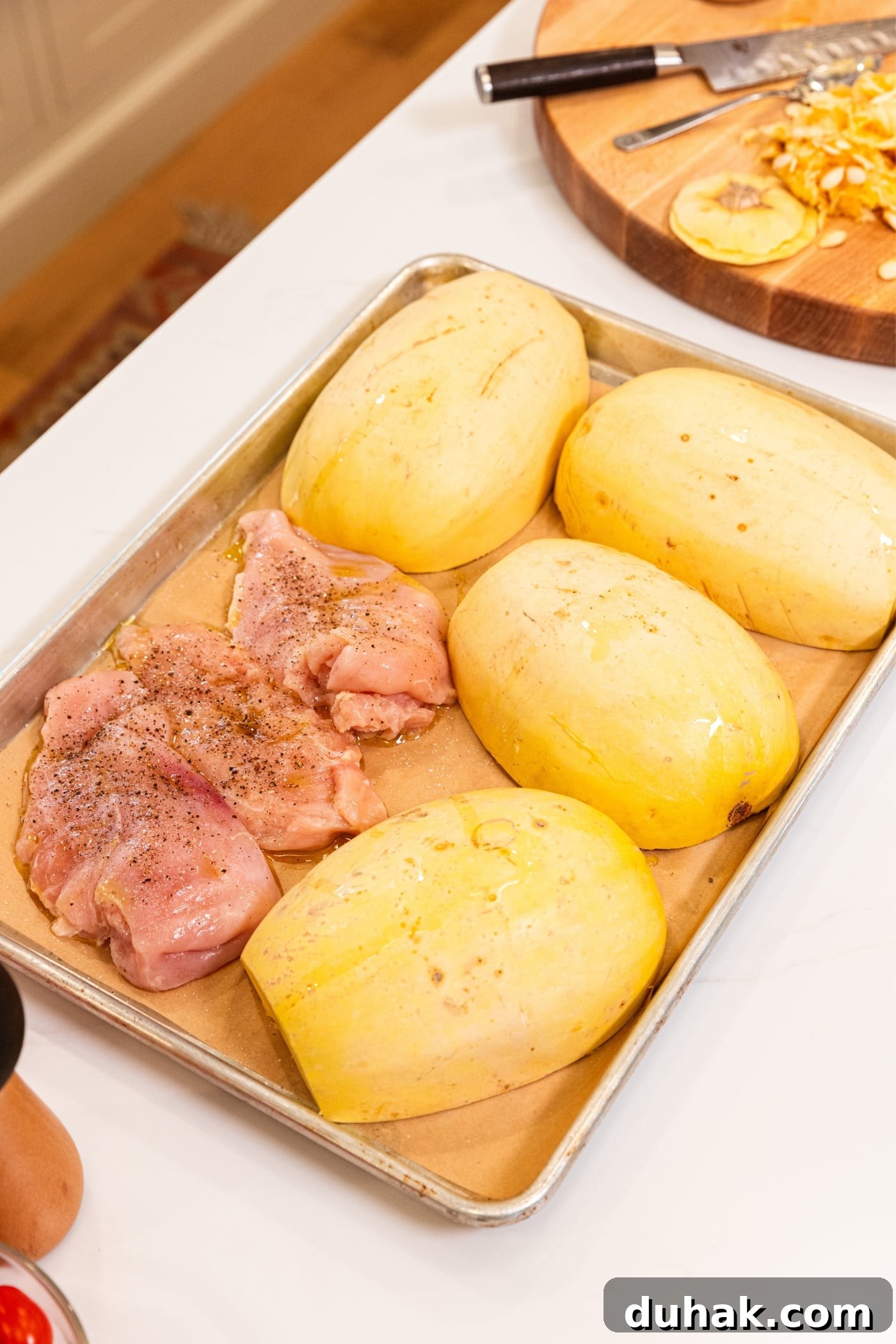 Cheesy Turkey & Spaghetti Squash Casserole Recipe 7 Large metal baking sheet with piece of parchment on bottom sitting on white surface with unbaked halves of spaghetti squash and raw chicken breasts with olive oil, salt, and pepper all over.