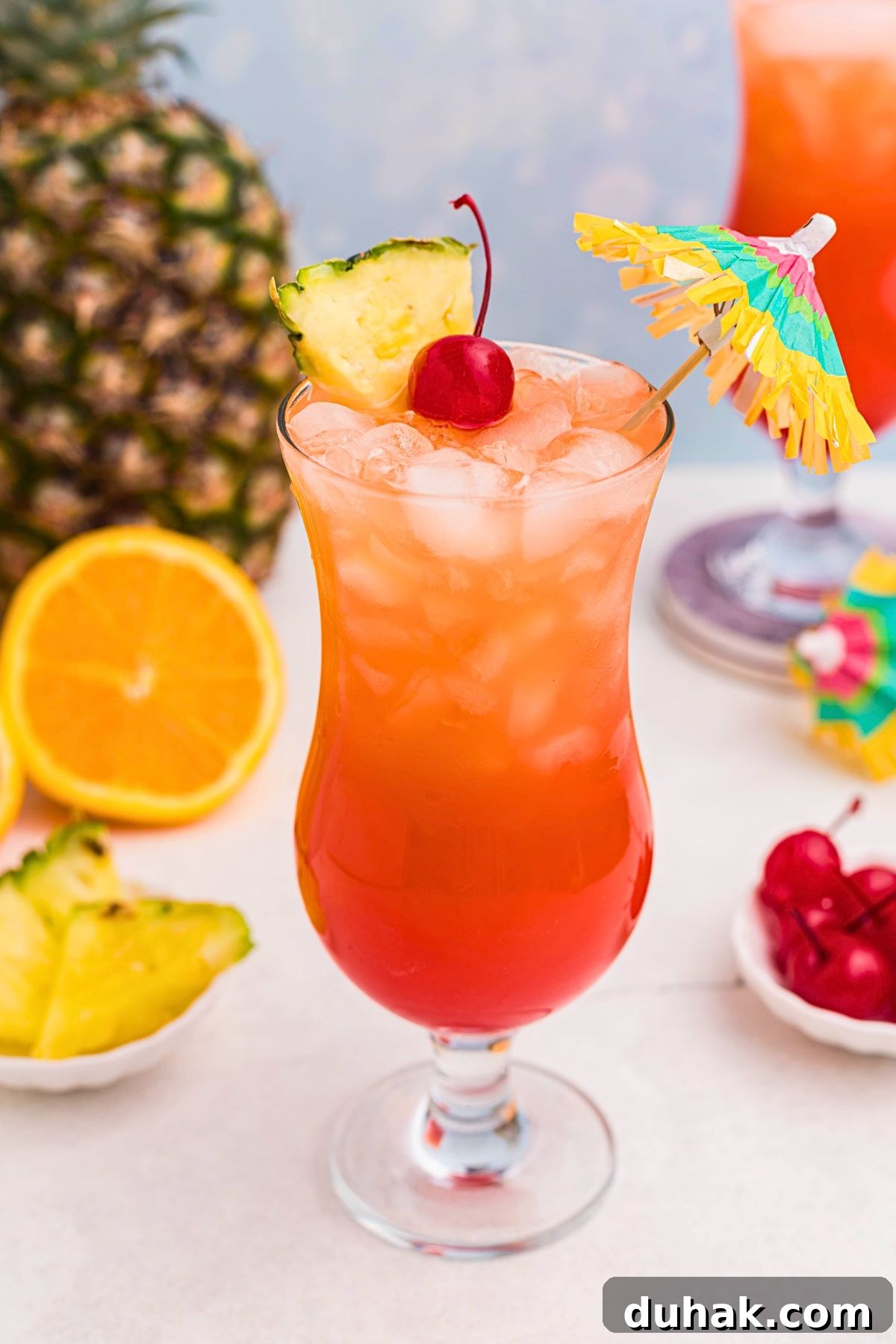 Bahama Mama Cocktail Recipe: Tropical Rum Drink Guide 2 Red and orange drink mixed together, with a paper umbrella, in a hurricane glass with orange slices and cherries on the marble table.