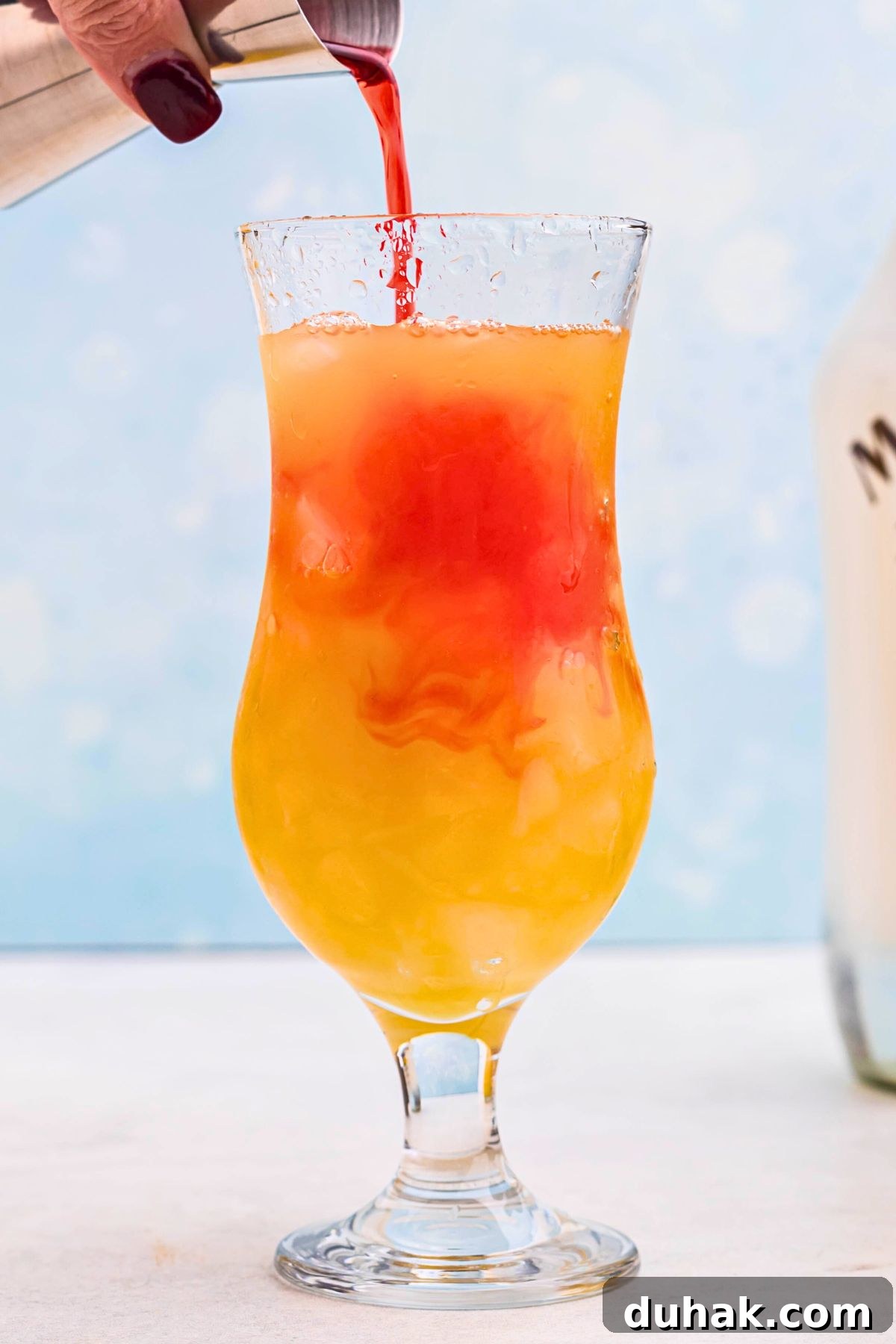 Bahama Mama Cocktail Recipe: Tropical Rum Drink Guide 6 Red grenadine being poured into orange drink in a hurricane glass.
