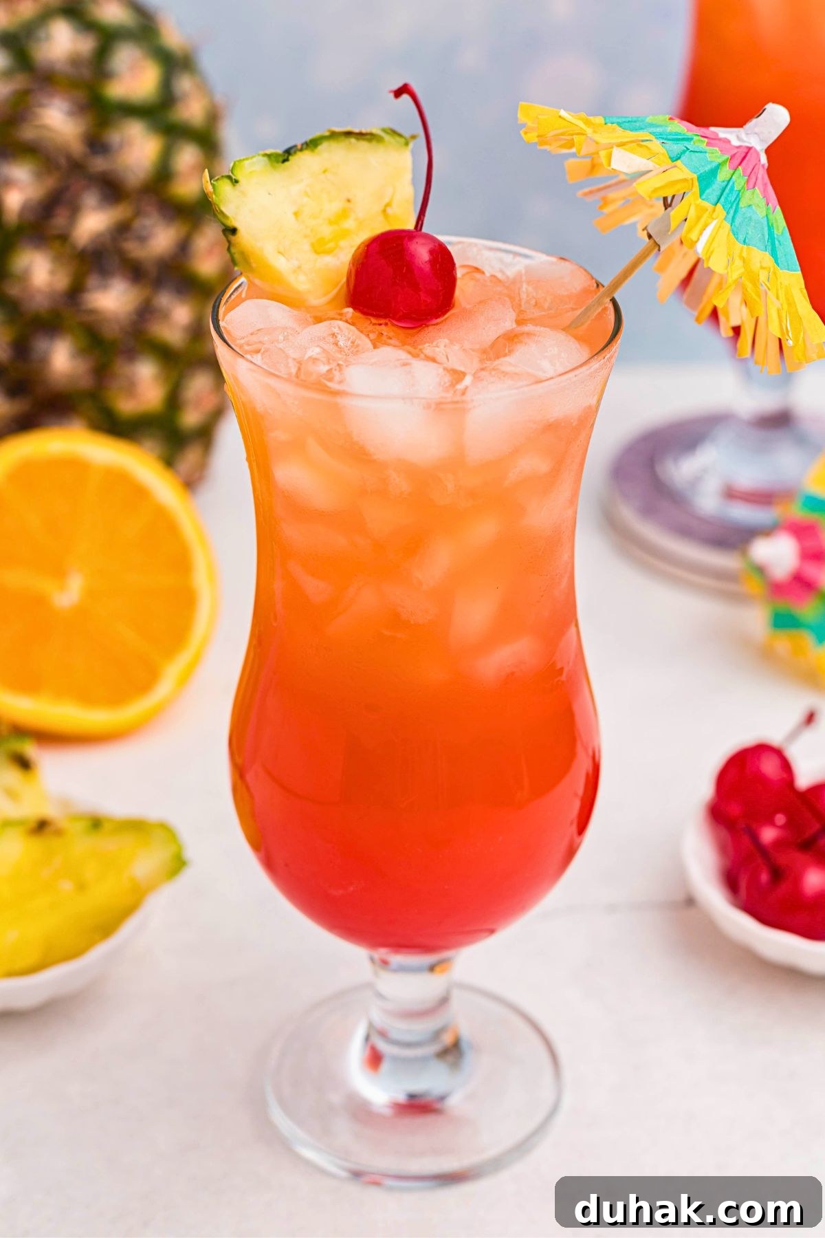 Bahama Mama Cocktail Recipe: Tropical Rum Drink Guide 8 Red and orange drink mixed together, with a paper umbrella, in a hurricane glass with orange slices and cherries on the marble table.
