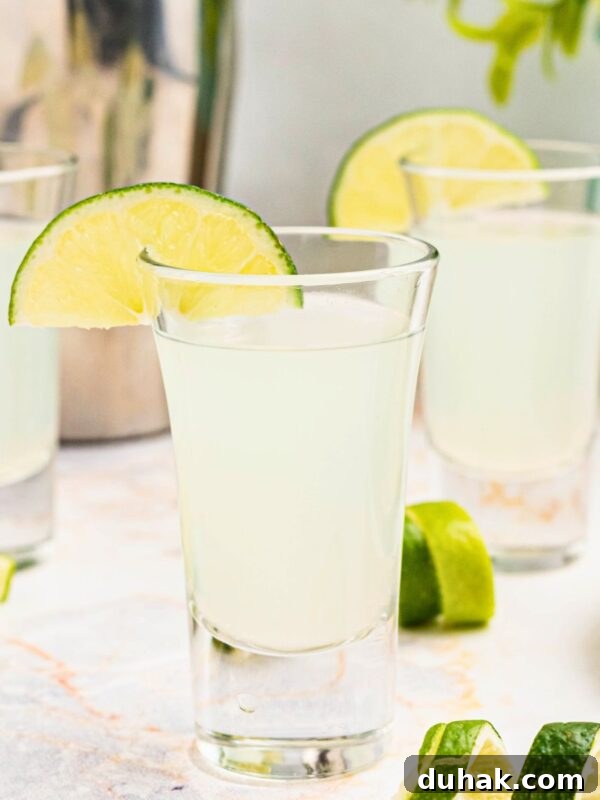 Bahama Mama Cocktail Recipe: Tropical Rum Drink Guide 9 Cold Kamikaze shots in shot glasses with lime wedges and lime curls on a marble table in front of a cocktail shaker.