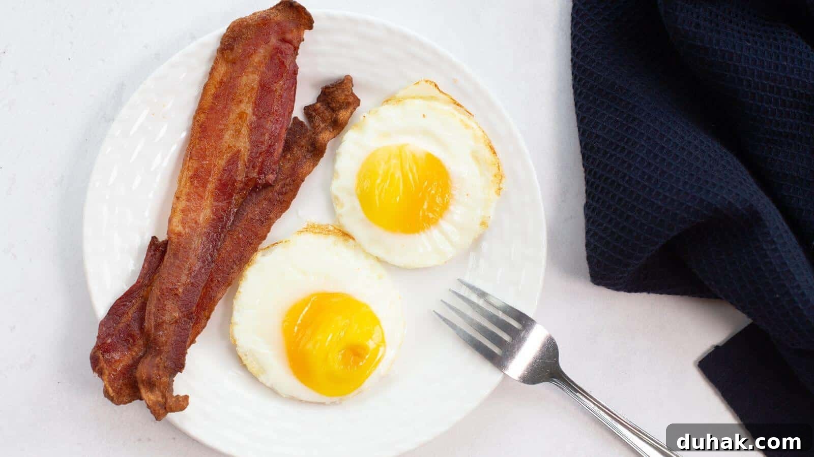 24 Low-Carb Morning Wins for Delicious, Satisfying Breakfasts 9 Plate with two sunny-side-up eggs, perfectly cooked like fried eggs in an air fryer, and two strips of crispy bacon, next to a fork and a dark cloth napkin.