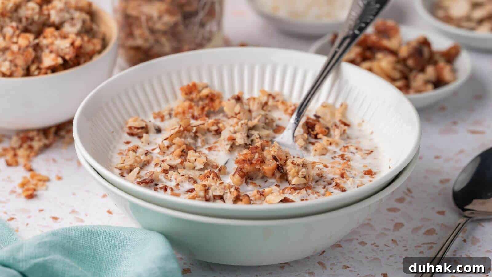 24 Low-Carb Morning Wins for Delicious, Satisfying Breakfasts 10 A bowl of granola mixed with milk, with a spoon inside. Other bowls of granola and toppings visible in the background. A teal cloth is partially visible on the side.
