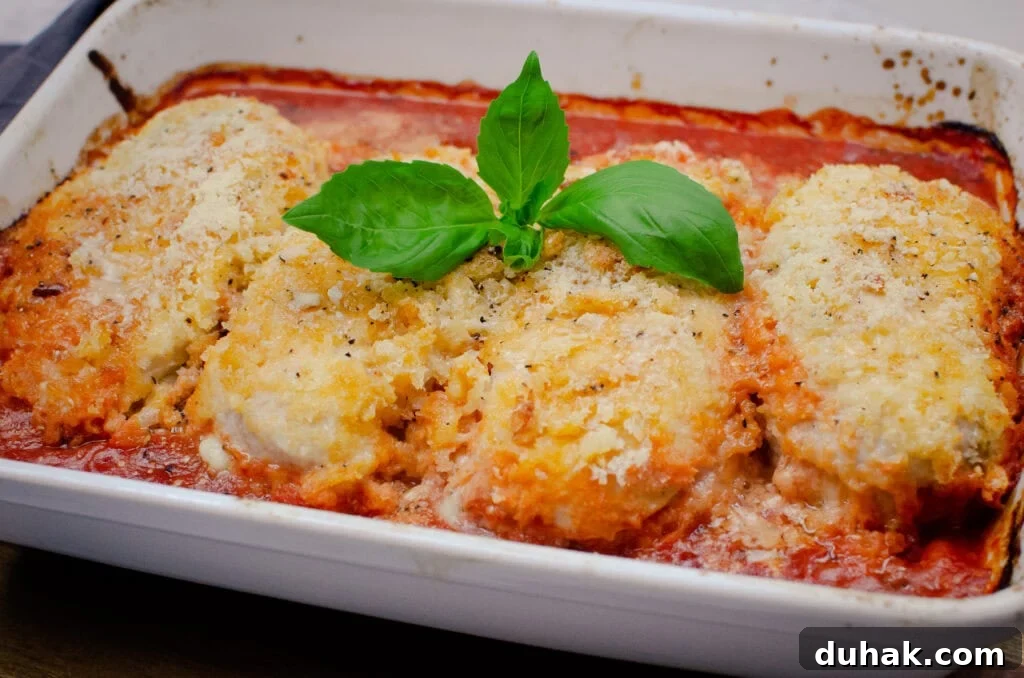 Four crispy chicken breasts with parmesan and fresh basil leaves on top in tomato sauce cooked in a white casserole dish