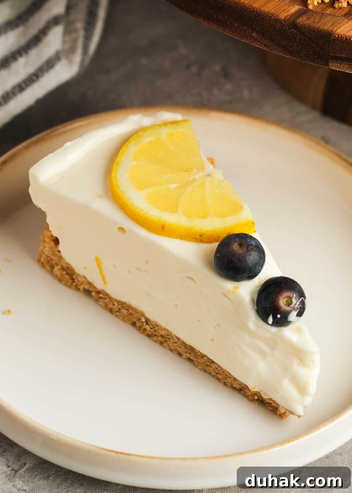 Lemon Cheesecake Recipe with Zesty Citrus Cream Filling 14 a white plate topped with a perfect slice of easy no-bake lemon cheesecake garnished with lemon slices and fresh blueberries