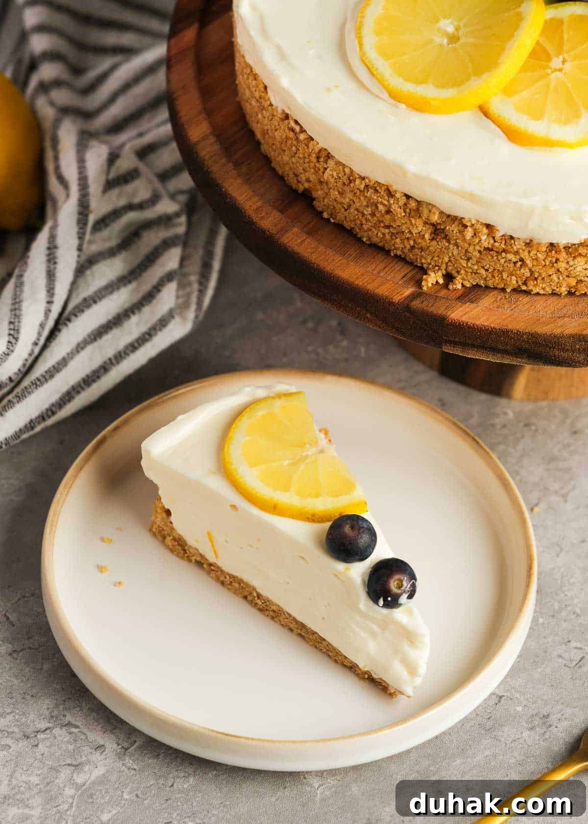 Lemon Cheesecake Recipe with Zesty Citrus Cream Filling 15 a white plate topped with a slice of no bake lemon cheesecake topped with a lemon slice and two blueberries; in the background there is the remaining cheesecake and a dish towel