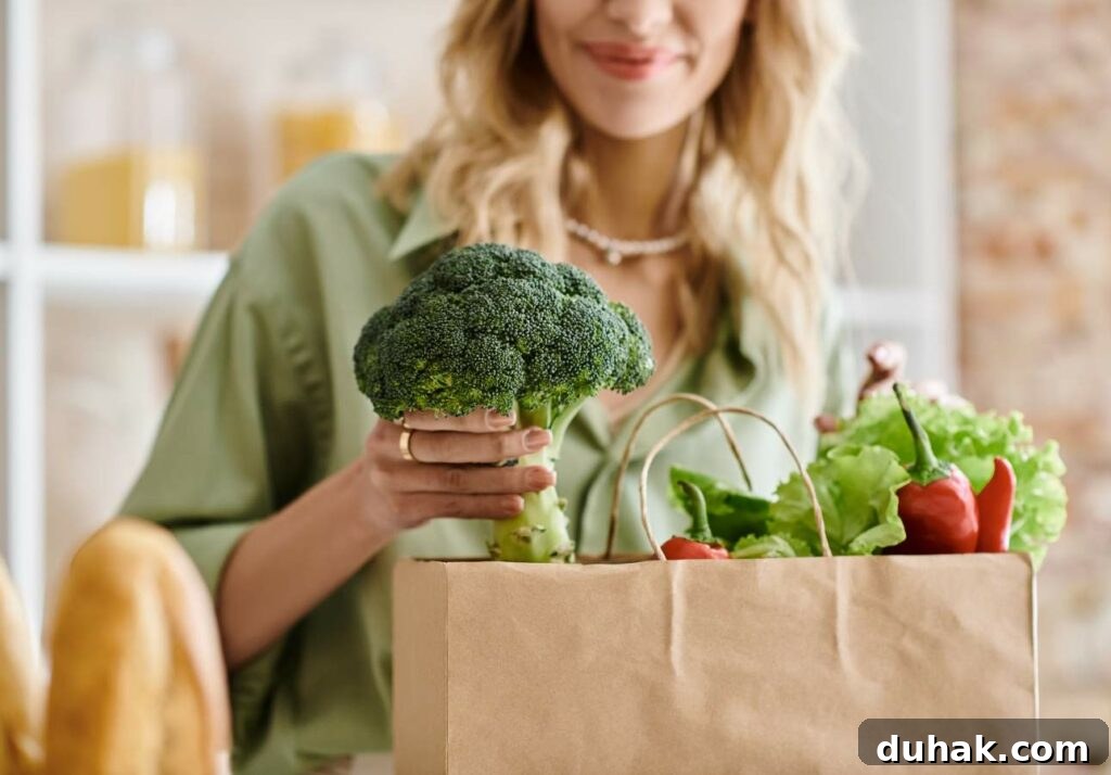 Low-Oxalate Leafy Greens to Add to Your Diet 2 A woman holding a broccoli and unpacking groceries from a brown paper bag filled with fresh vegetables like lettuce and bell peppers in a kitchen.