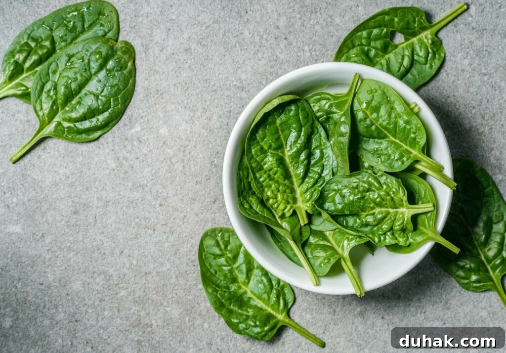 Low-Oxalate Leafy Greens to Add to Your Diet 3 A white bowl filled with fresh spinach leaves, placed on a gray surface with a few spinach leaves scattered around.