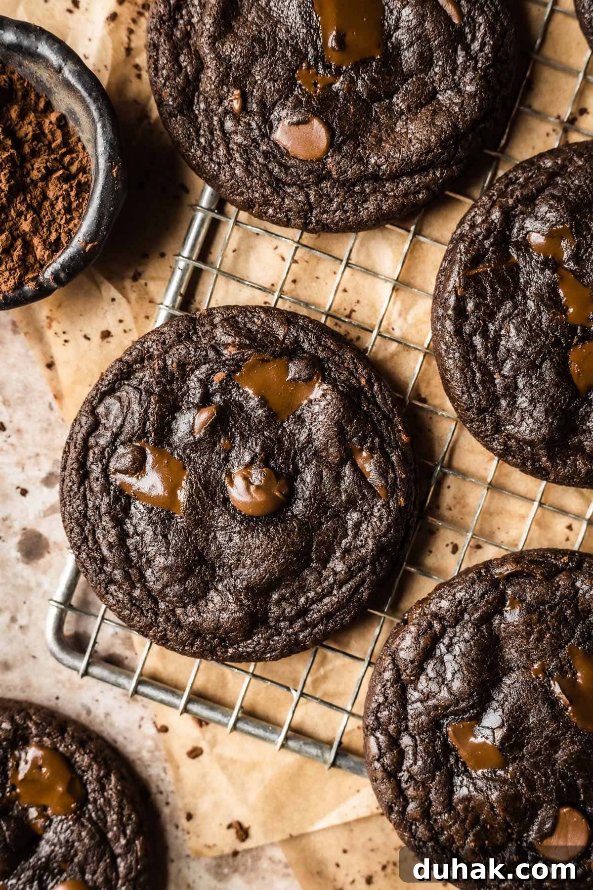 Double Cocoa Chocolate Cookies: Rich, Soft Baked Treats 2 A metal cooling rack with dark chocolate cookies on top of brown parchment paper with a small bowl of cocoa powder nearby.