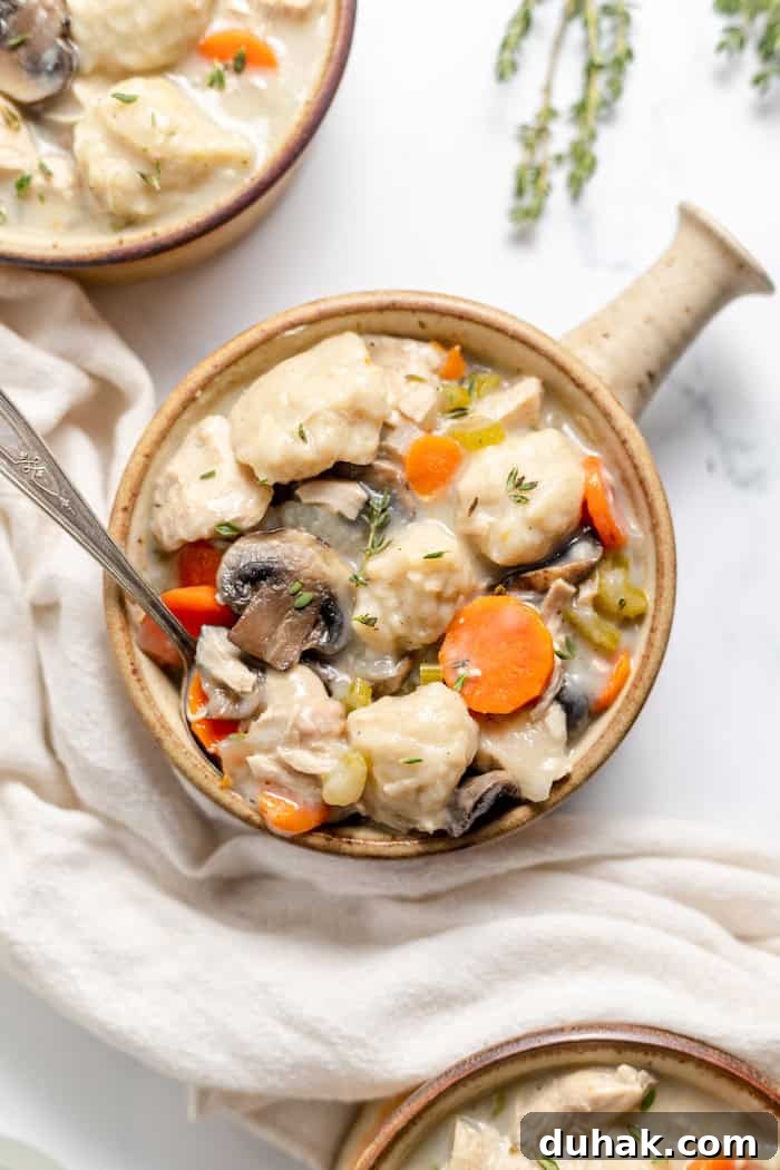 Comforting Gluten-Free Chicken with Fluffy Dumplings 4 Image, taken from above, shows a small ceramic crock of chicken and dumplings soup on a white counter top with a spoon in it.