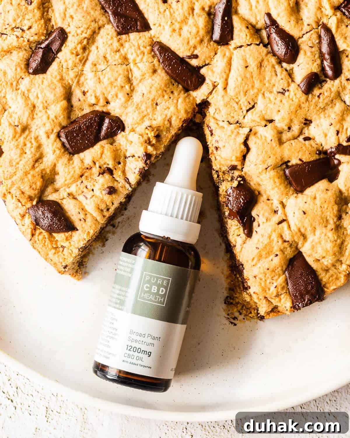 A Giant Chocolate Chip Cookie on a white plate, a slice is missing and in its place is a bottle of cbd oil.