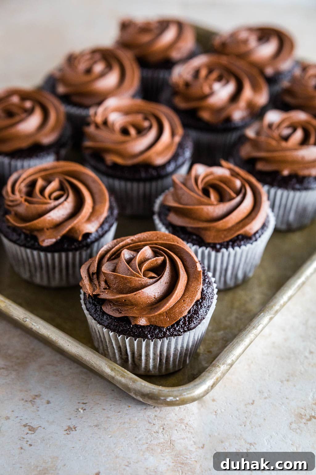 A tray of gluten-free chocolate cupcakes with rich, swirled chocolate frosting on top, arranged in neat rows on a metal baking sheet.