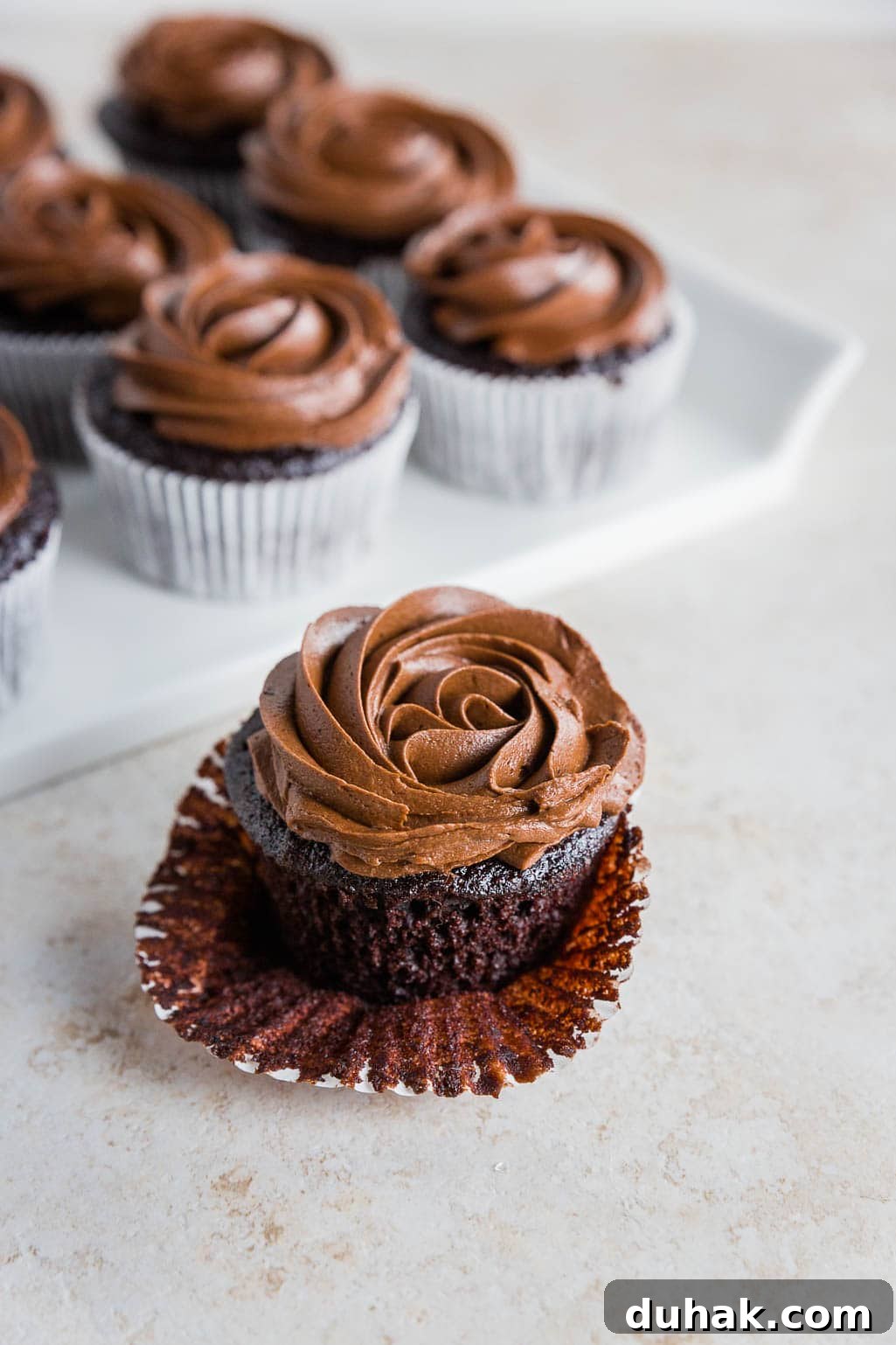 A gluten-free chocolate cupcake with swirled chocolate frosting sits in an open paper liner in the foreground, with more similarly decorated gluten-free chocolate cupcakes on a white tray in the background.