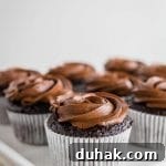 A close-up of several gluten-free chocolate cupcakes in white paper liners, topped with swirls of chocolate frosting, arranged on a white tray against a light background.