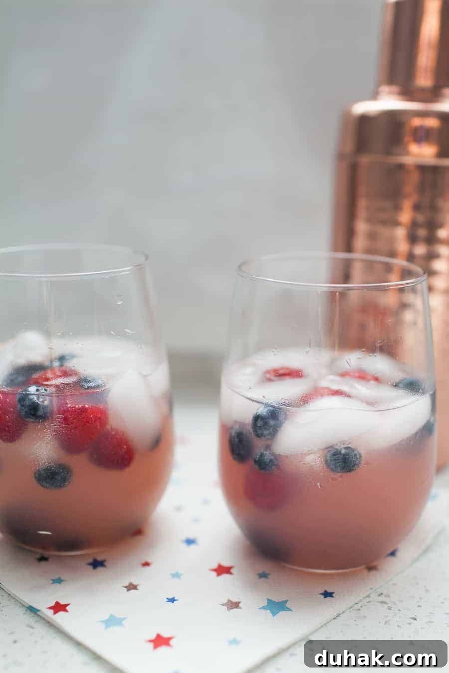Classic Tequila Soda Recipe: Crisp Low-Calorie Cocktail Guide 2 Two glasses of a refreshing pink beverage, resembling a White Wine Spritzer, with ice cubes and mixed berries on a star-patterned napkin. A copper cocktail shaker is in the background.