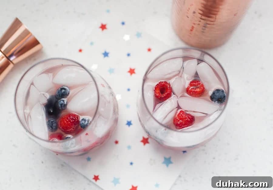 Classic Tequila Soda Recipe: Crisp Low-Calorie Cocktail Guide 3 Two glasses of White Wine Spritzer with raspberries and blueberries sit on a star-patterned napkin, alongside a copper cocktail shaker and jigger on a white surface.