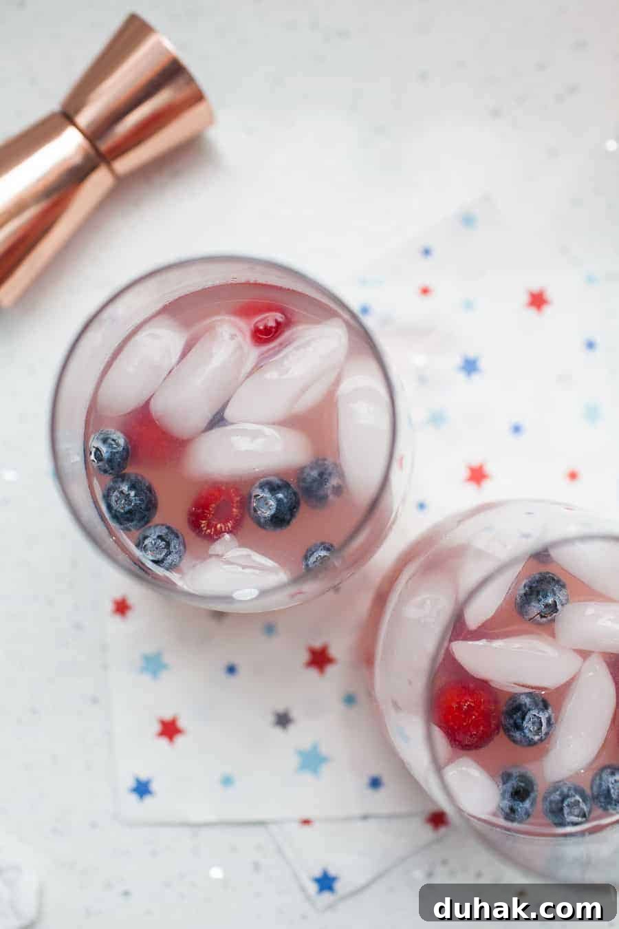Classic Tequila Soda Recipe: Crisp Low-Calorie Cocktail Guide 5 Two glasses of a refreshing White Wine Spritzer with ice, blueberries, and raspberries sit on a white surface with a star-printed napkin. A copper jigger is visible at the top left corner, hinting at the careful crafting of these delightful drinks.